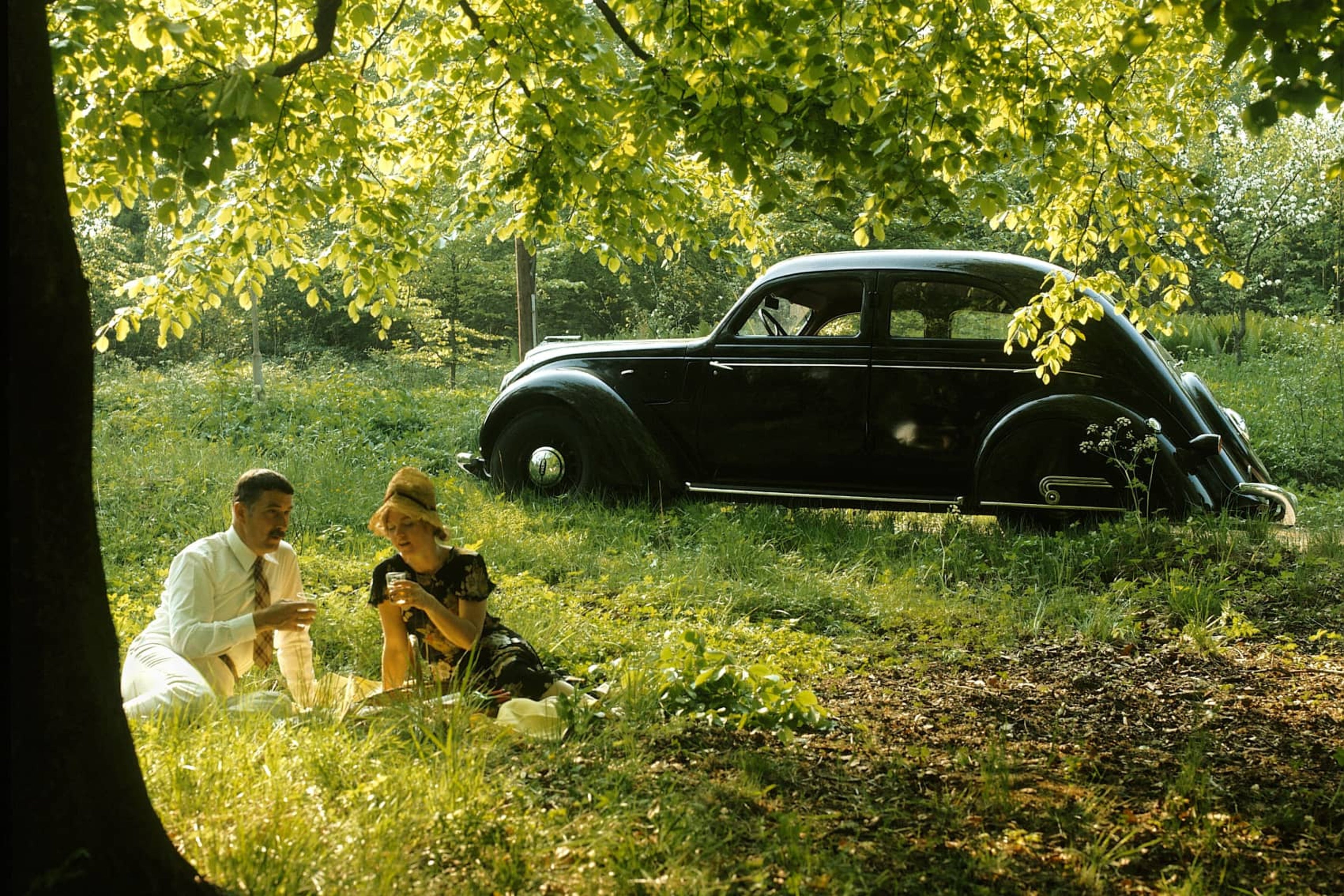 Un Volvo PV36 Carioca aparcado en un prado, con dos personas haciendo un picnic delante del coche.