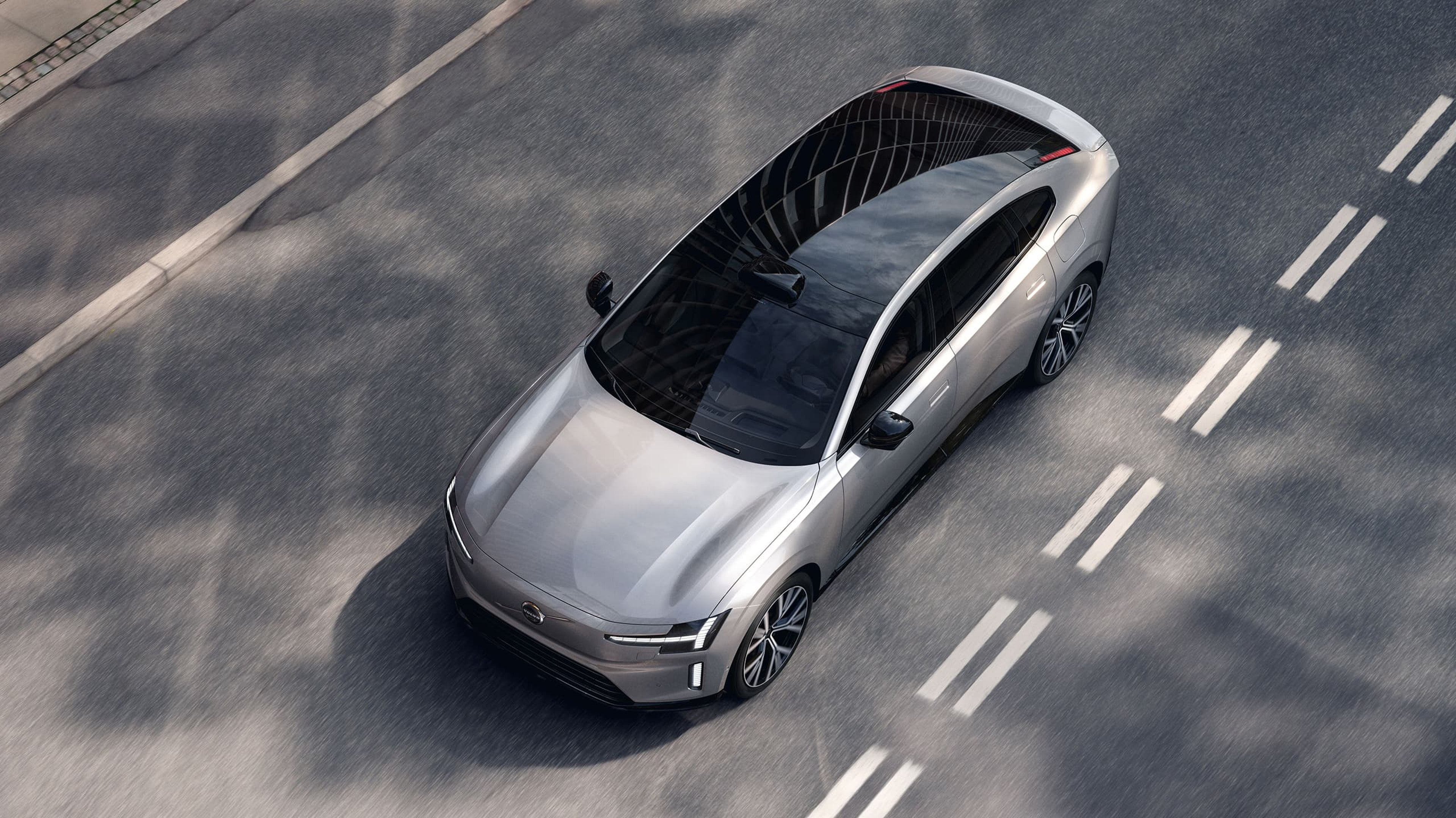 Vue de dessus d'une Volvo ES90 argentée roulant sur une route, mettant en valeur sa forme aérodynamique et son toit panoramique noir.