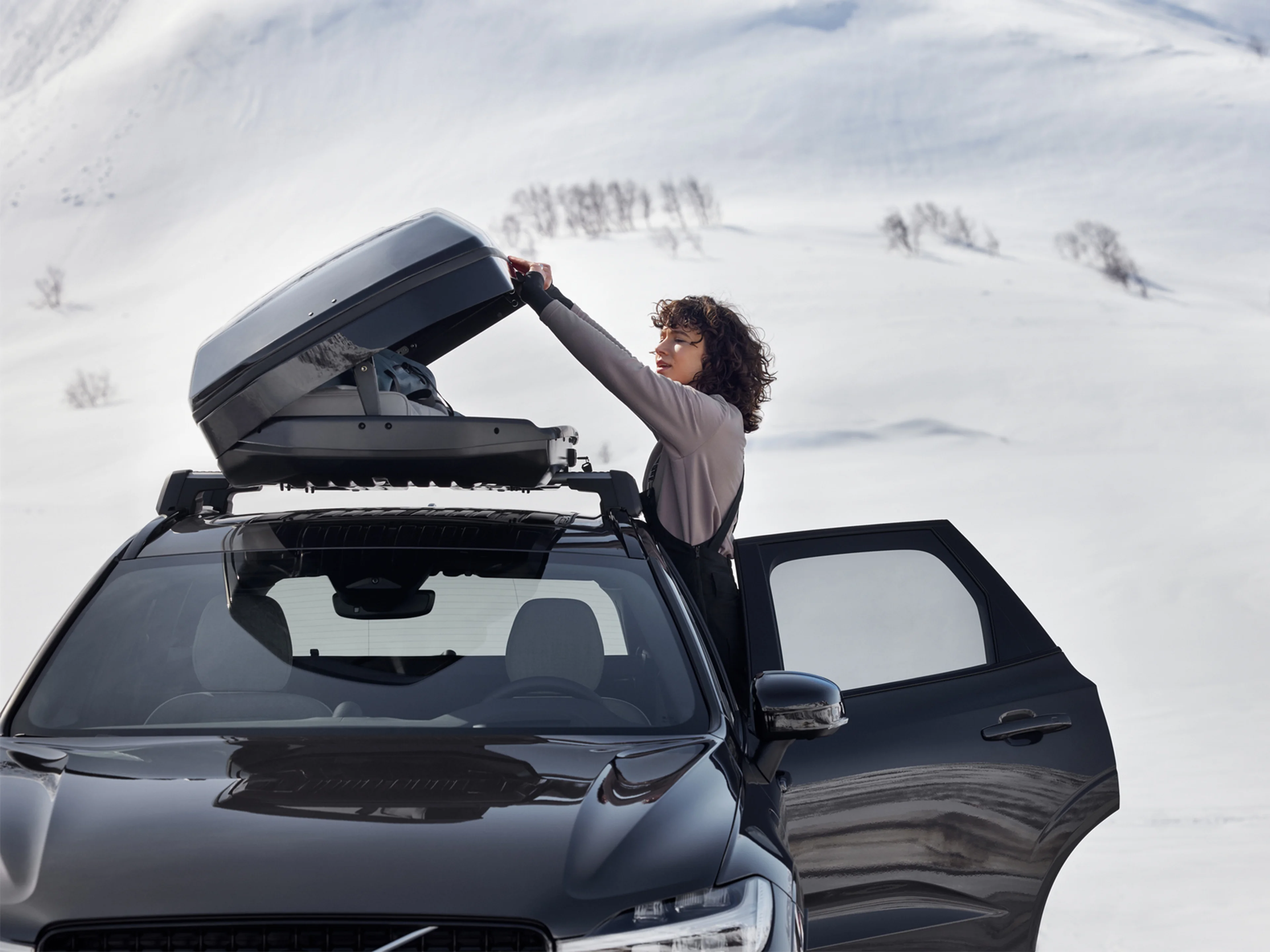 Nainen avaa Platinum Grey -värisen Volvo XC60 auton katolla olevaa kattolaatikkoa lumisessa maisemassa.