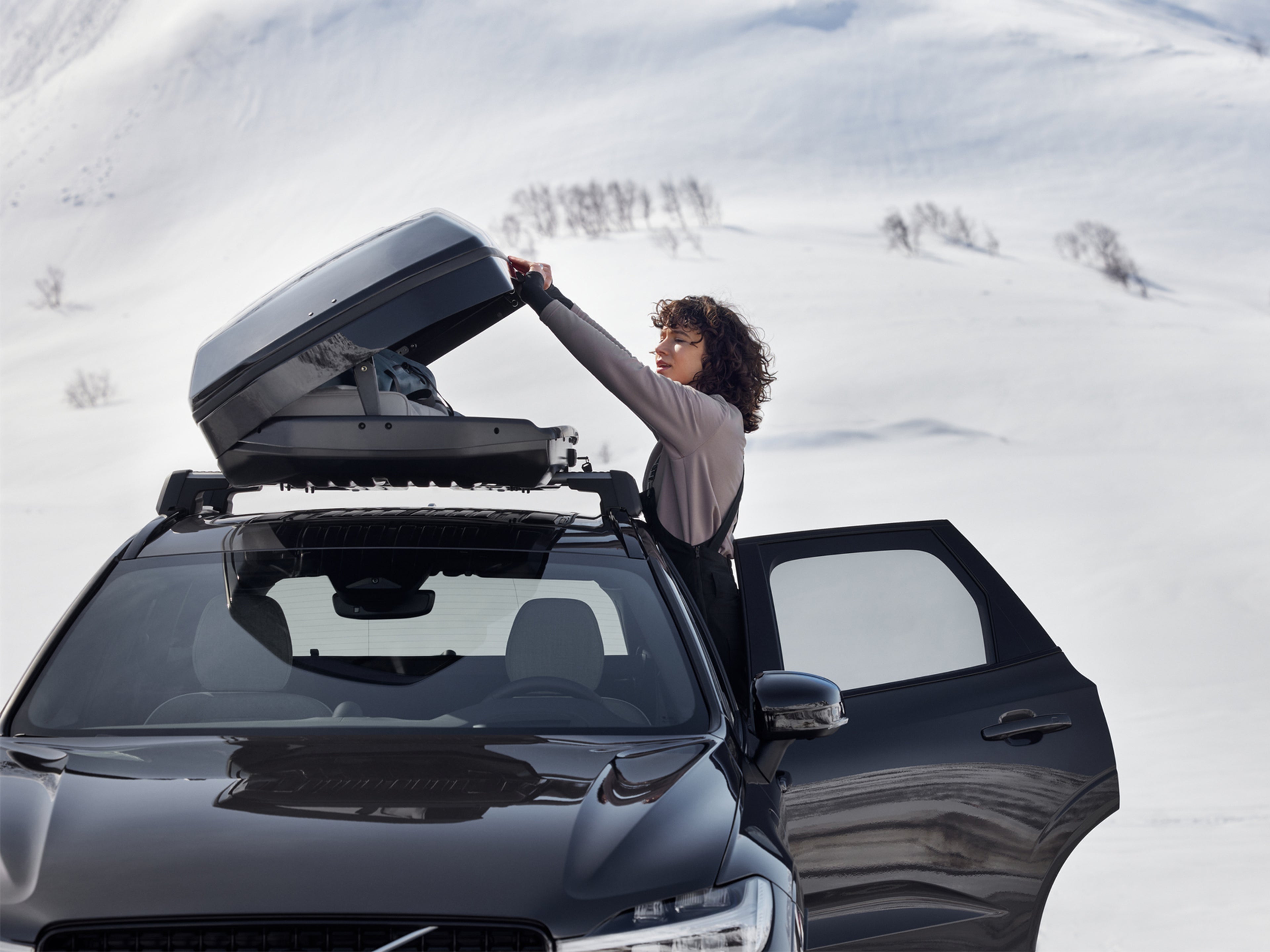 Nainen avaa Platinum Grey -värisen Volvo XC60 auton katolla olevaa kattolaatikkoa lumisessa maisemassa.