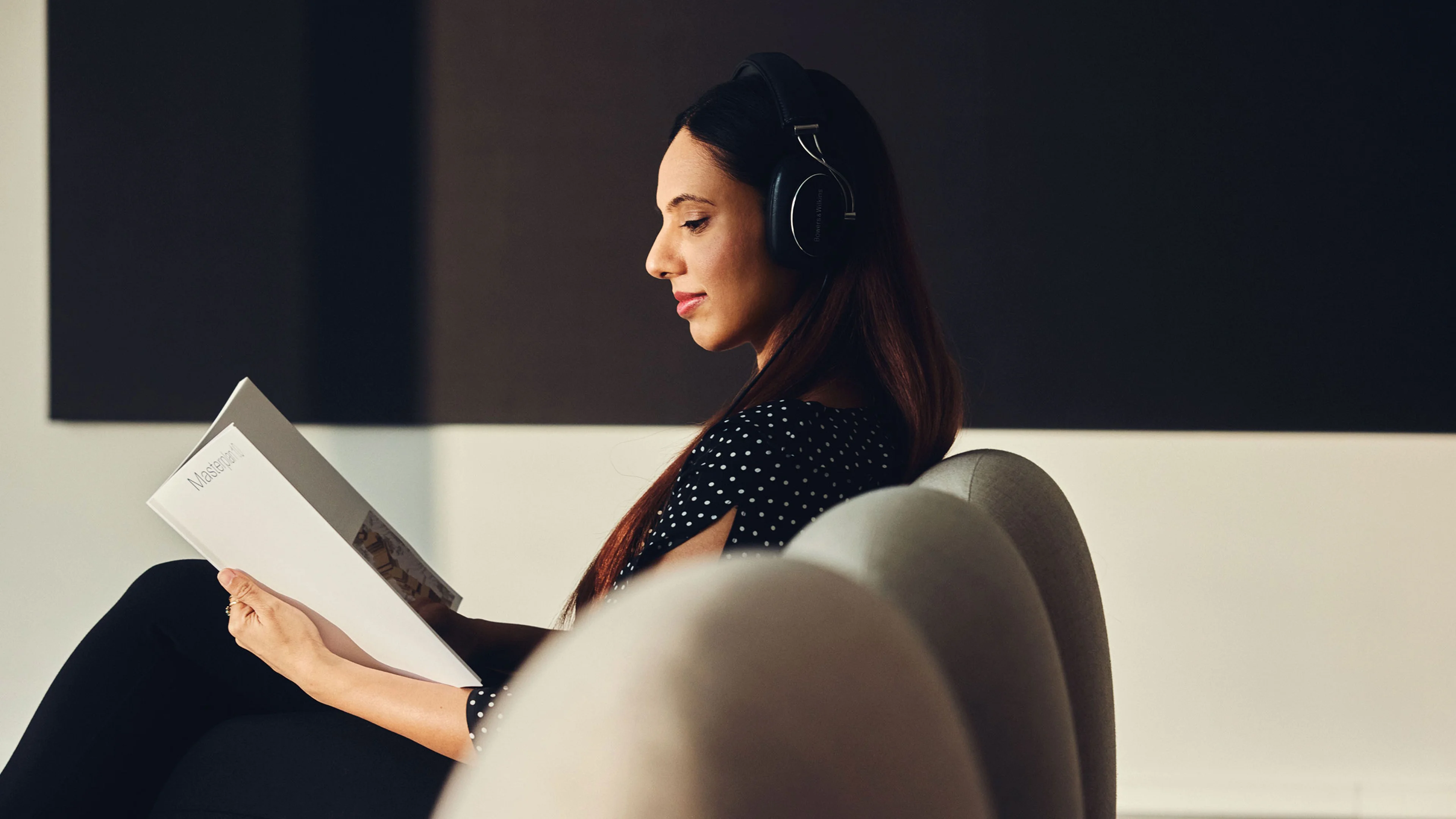 Eine Frau hat Kopfhörer auf und sitzt mit einer Broschüre auf einem Sofa. Sie liest die Broschüre.