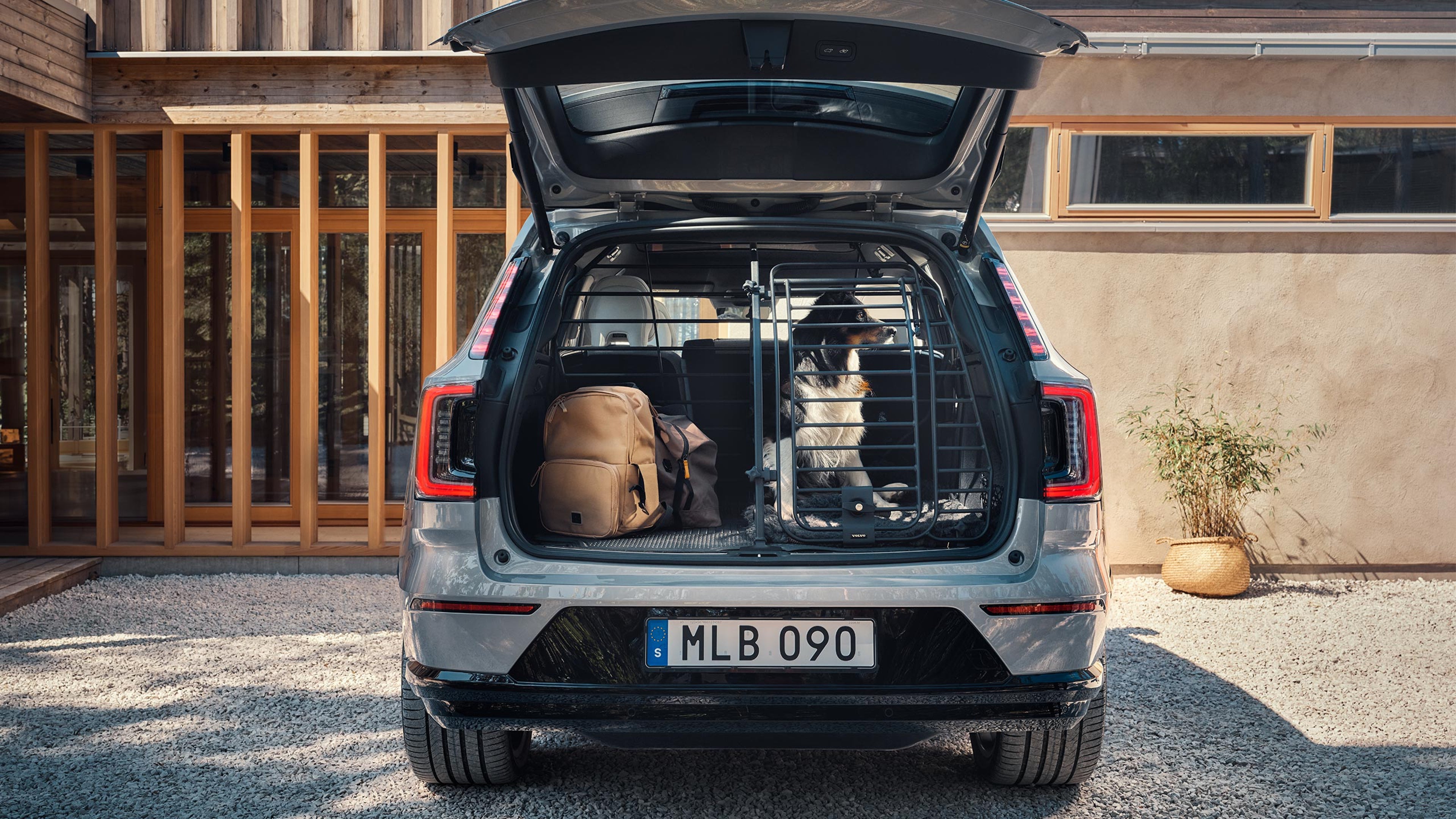 Vapour grey -värinen Volvo EX90 täyssähköinen katumaasturi pysäköitynä asuintalon pihaan. Auton takaluukku on auki ja takatilaan on asennettu Volvon koiraportti lisävaruste. Portin takana istuu mustavalkoinen, keskikokoinen koira. Jäljelle jäävään tyhjään tilaan on nostettu kasseja.