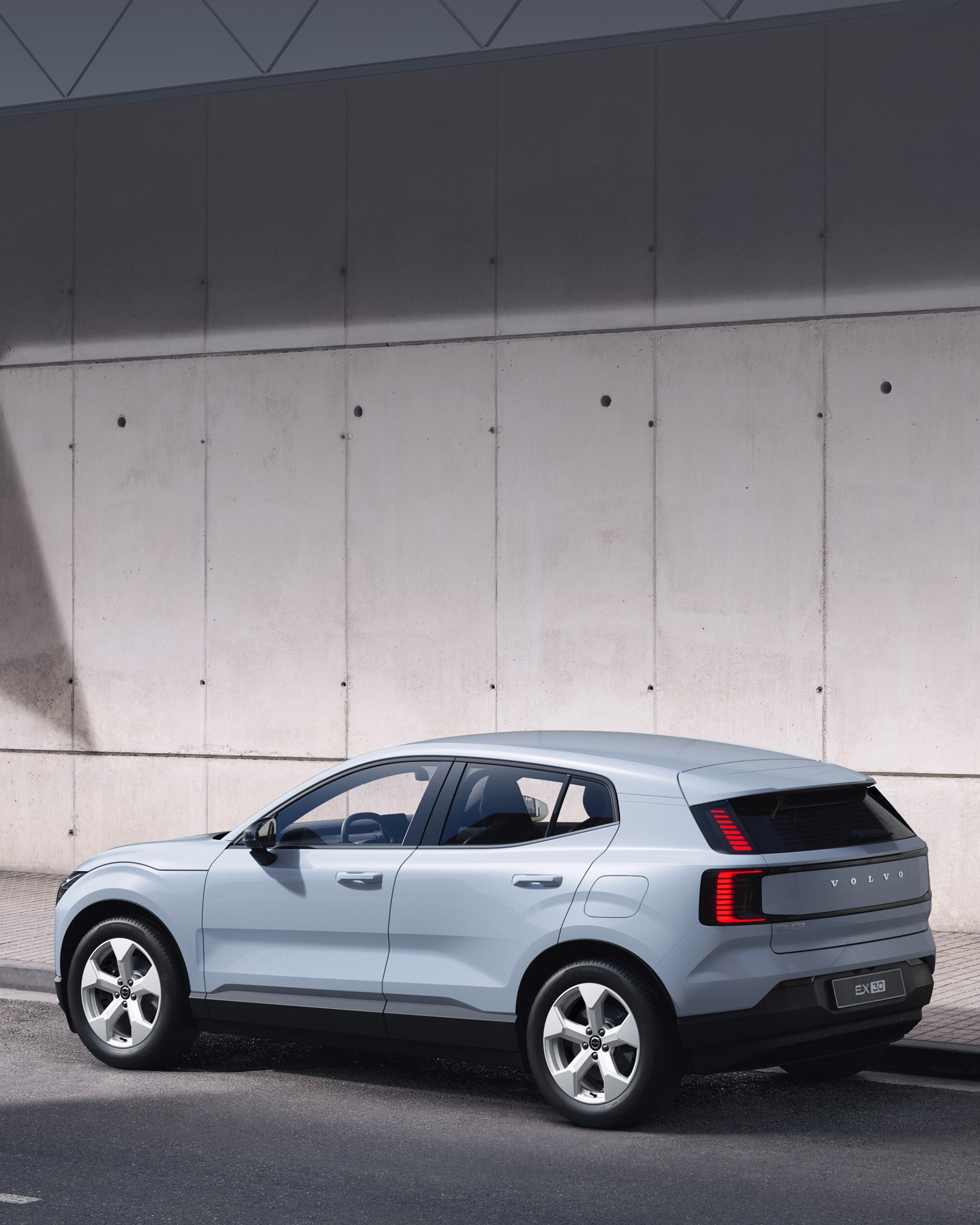 Cloud blue -värinen Volvo EX30 Essential täyssähköinen katumaasturi pysäköitynä tien varteen. Taustalla betoniseinä.