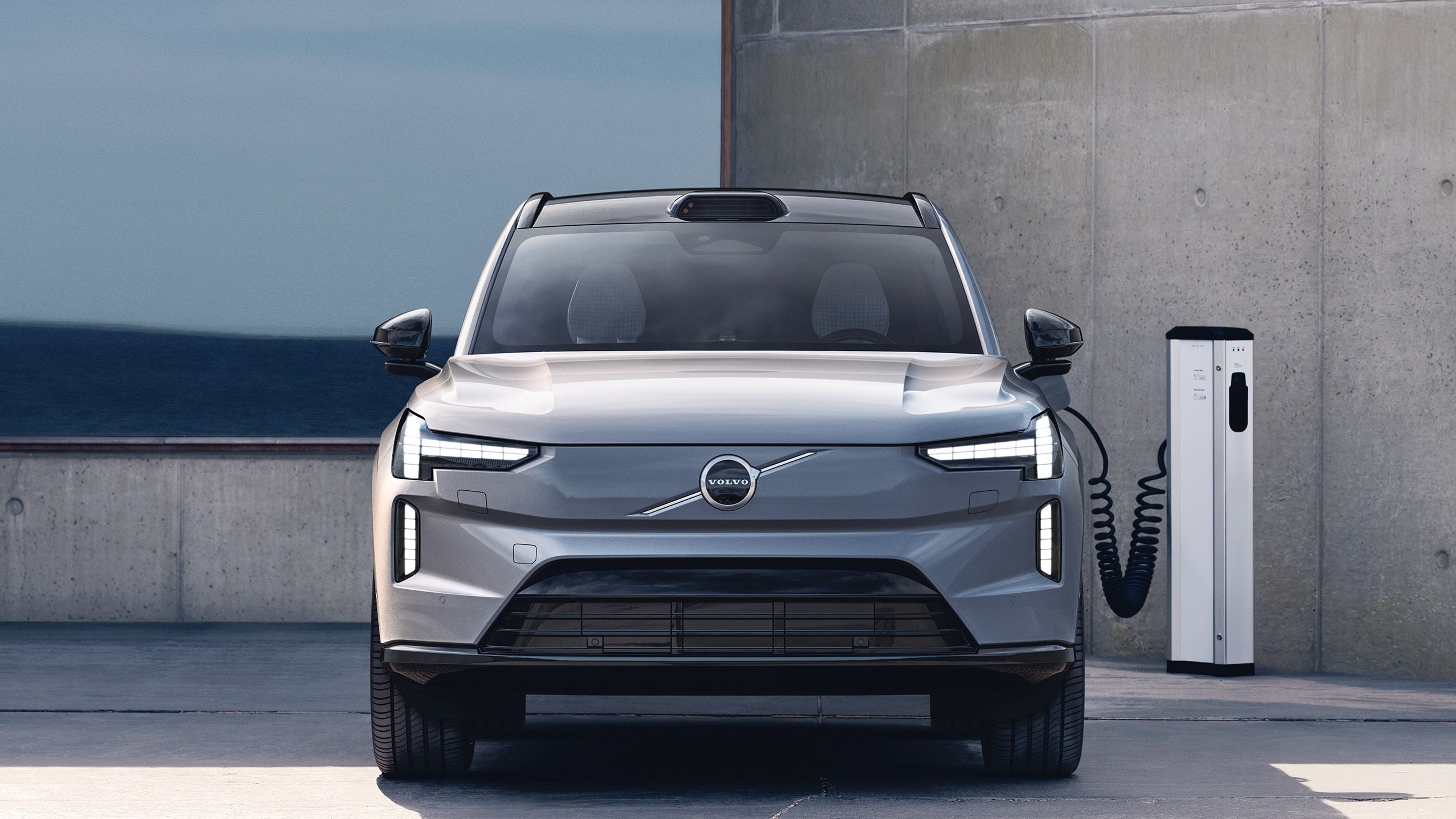 Vapour grey -värinen Volvo EX90 täyssähköinen katumaasturi kytkettynä latauspisteeseen betonirakennuksen edustalla.
