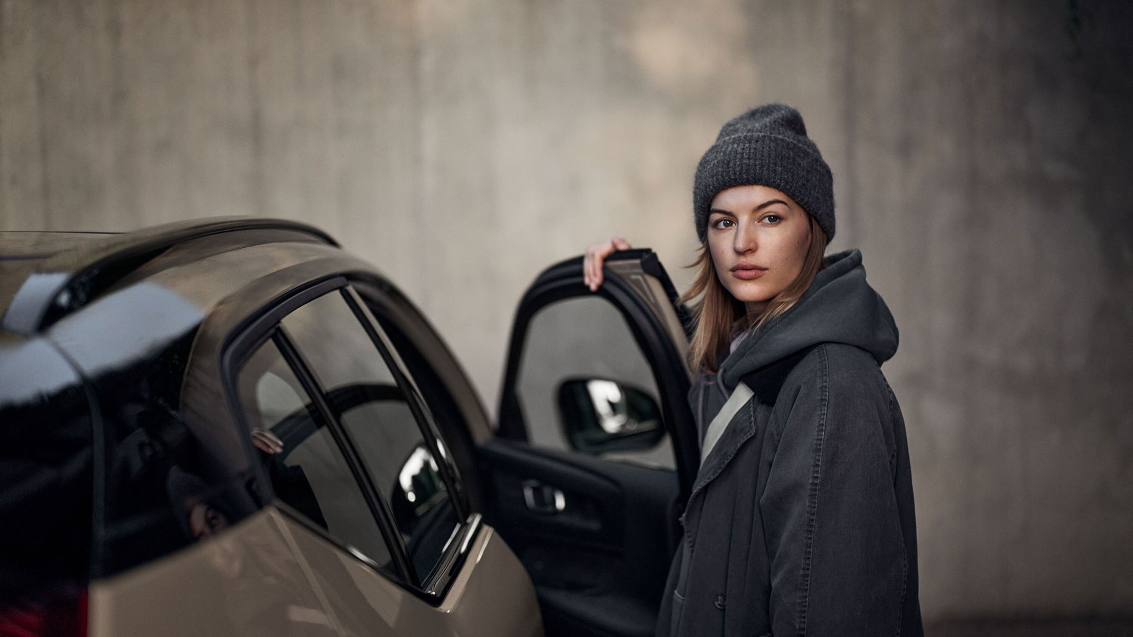 Vrouw in een grijze winterjas en muts die het portier van een bronskleurige Volvo EX40 opent.
