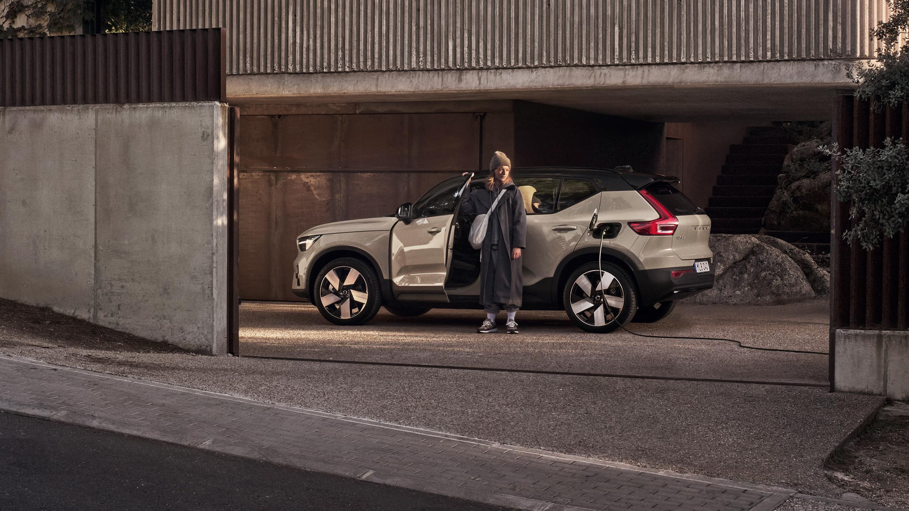Rennosti pukeutunut nainen seisoo betonitalon pihaan pysäköidyn ja lataukseen kytketyn Sand dune -värisen Volvo EX40 täyssähköisen katumaasturin kuljettajanpuoleisen oven ja auton välissä ja katselee pihaa.