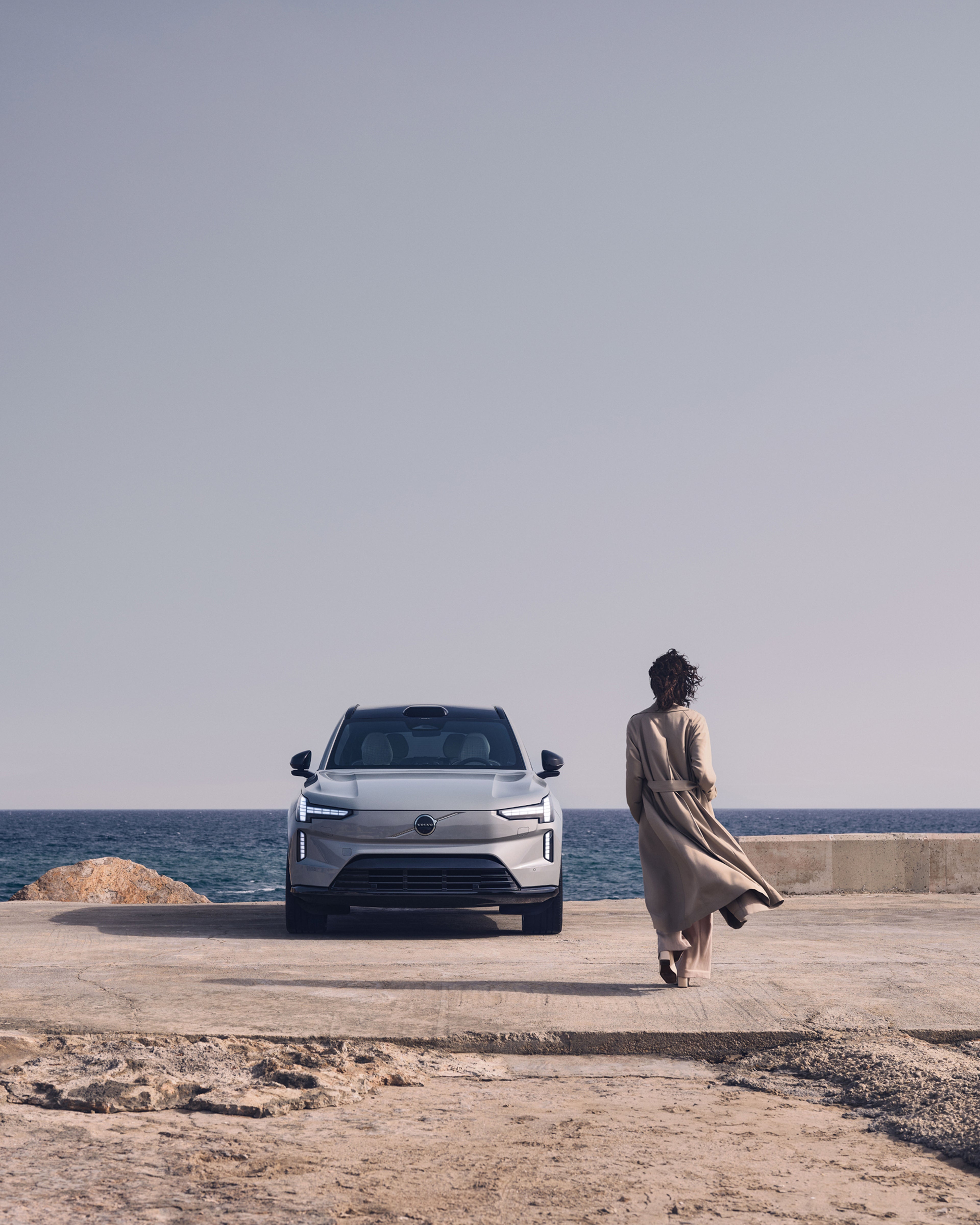 Femme marchant vers une EX90 garée au bord d’une route en bord de mer.