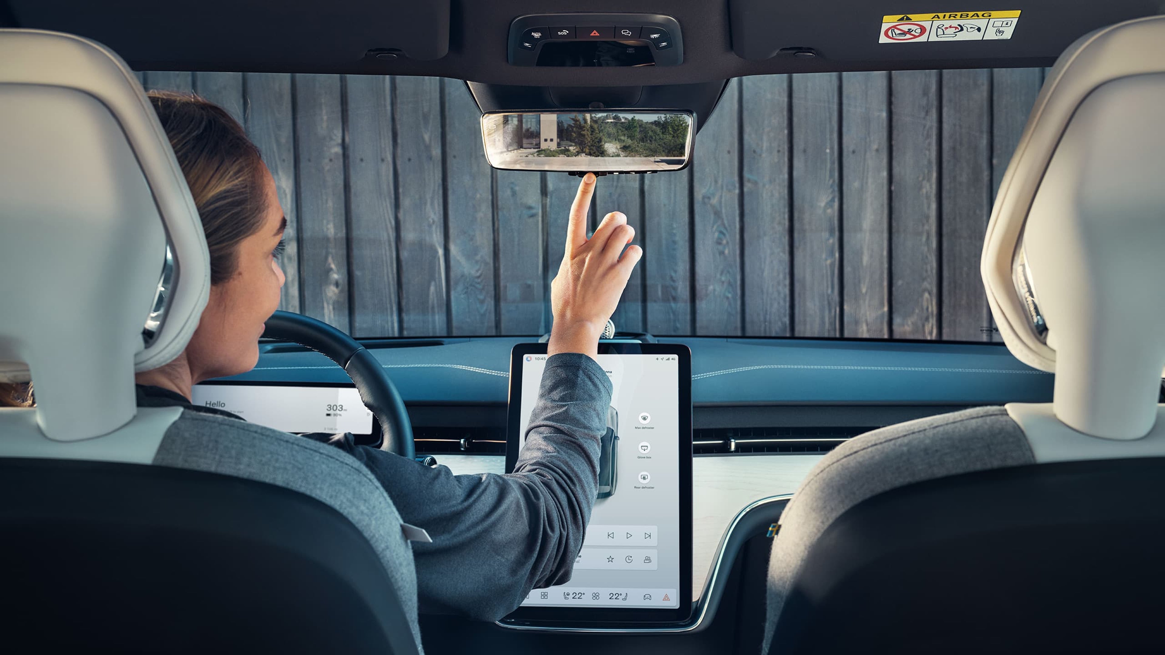 Intérieur de la Volvo EX90, vu depuis la banquette arrière. Une femme assise sur le siège conducteur ajuste le rétroviseur numérique.