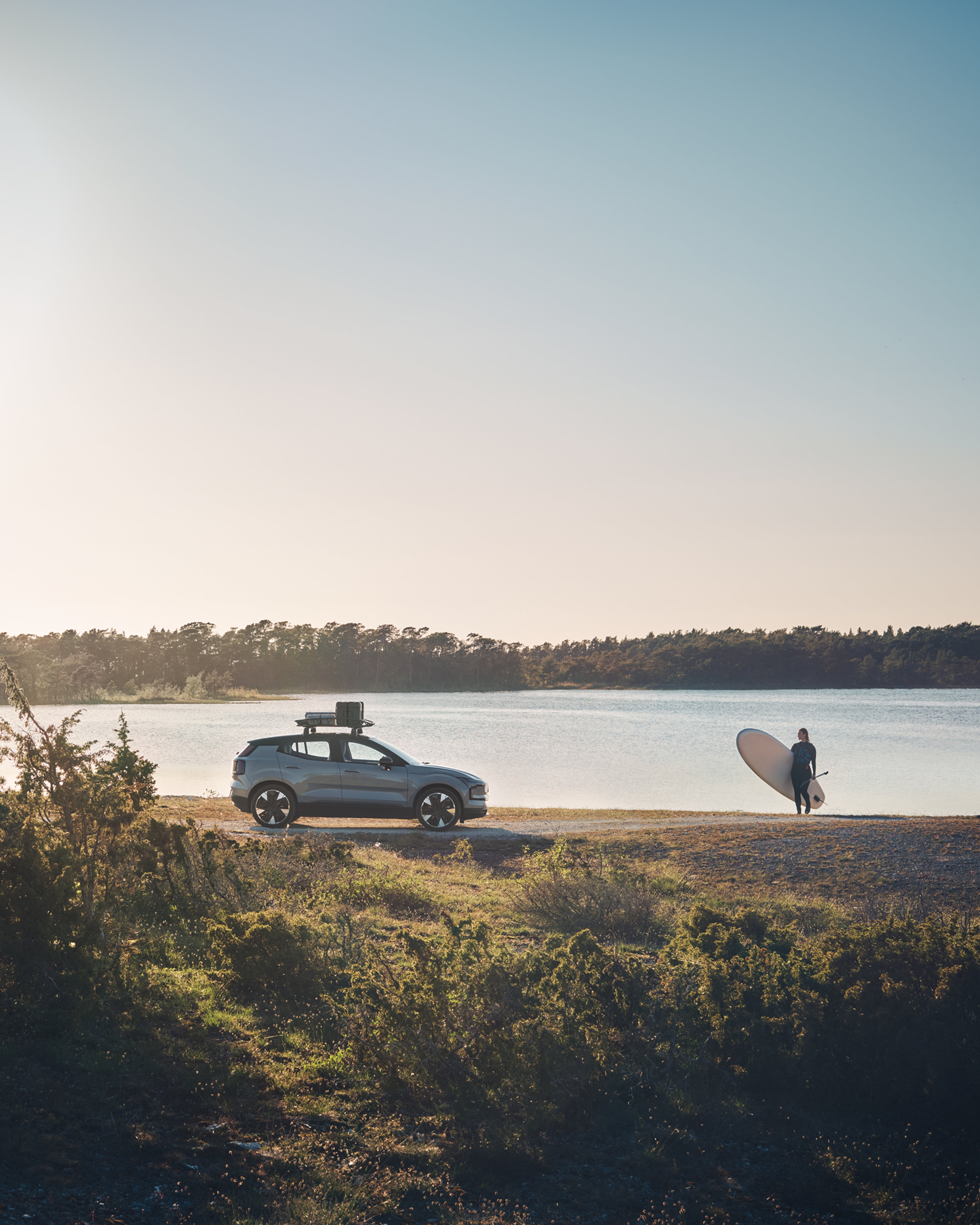 Side view of the Volvo EX30 with a woman holding a surf board next to the water.