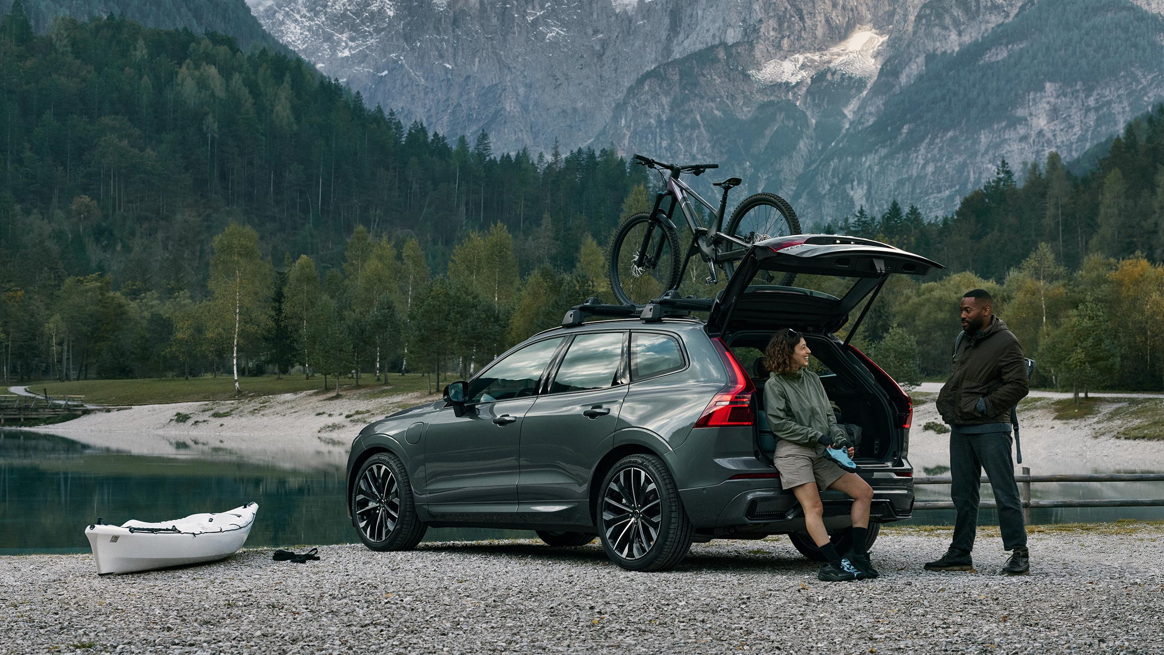 Vuoriston laaksojärven rantaan pysäköidyn Forest lake -värisen Volvo XC60 lataushybridikatumaasturin takatilan reunalla istuu retkeilijänainen, joka juttelee auton vieressä seisovan miehen kanssa. Auton katolle on kiinnitetty taakkatelineisiin polkupyörä ja auton vasemmalla puolella maassa on valkoinen kajakki.