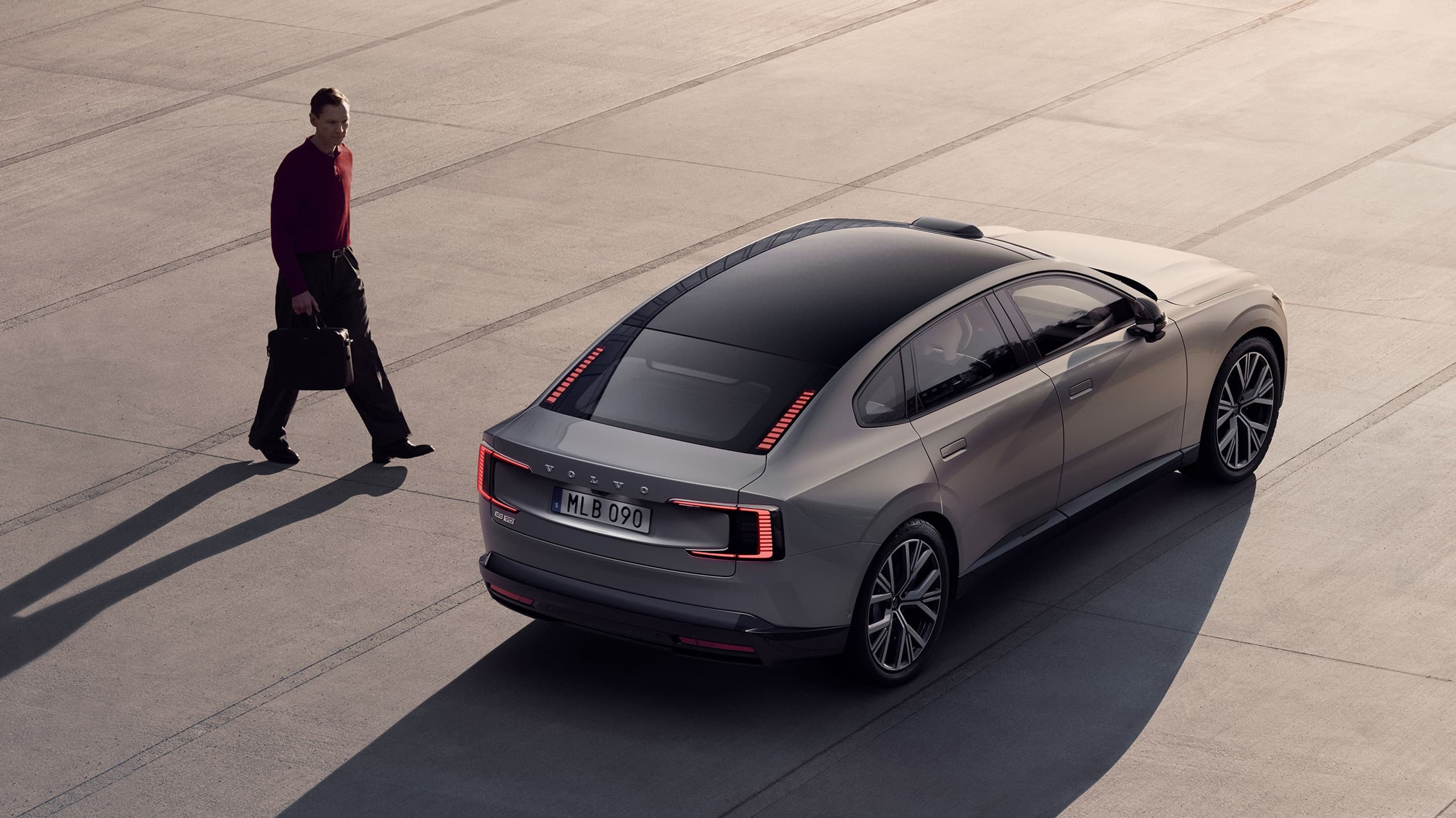 Vue arrière de la Volvo ES90 argentée sur une surface en béton, avec un homme tenant une mallette marchant à côté de la voiture.