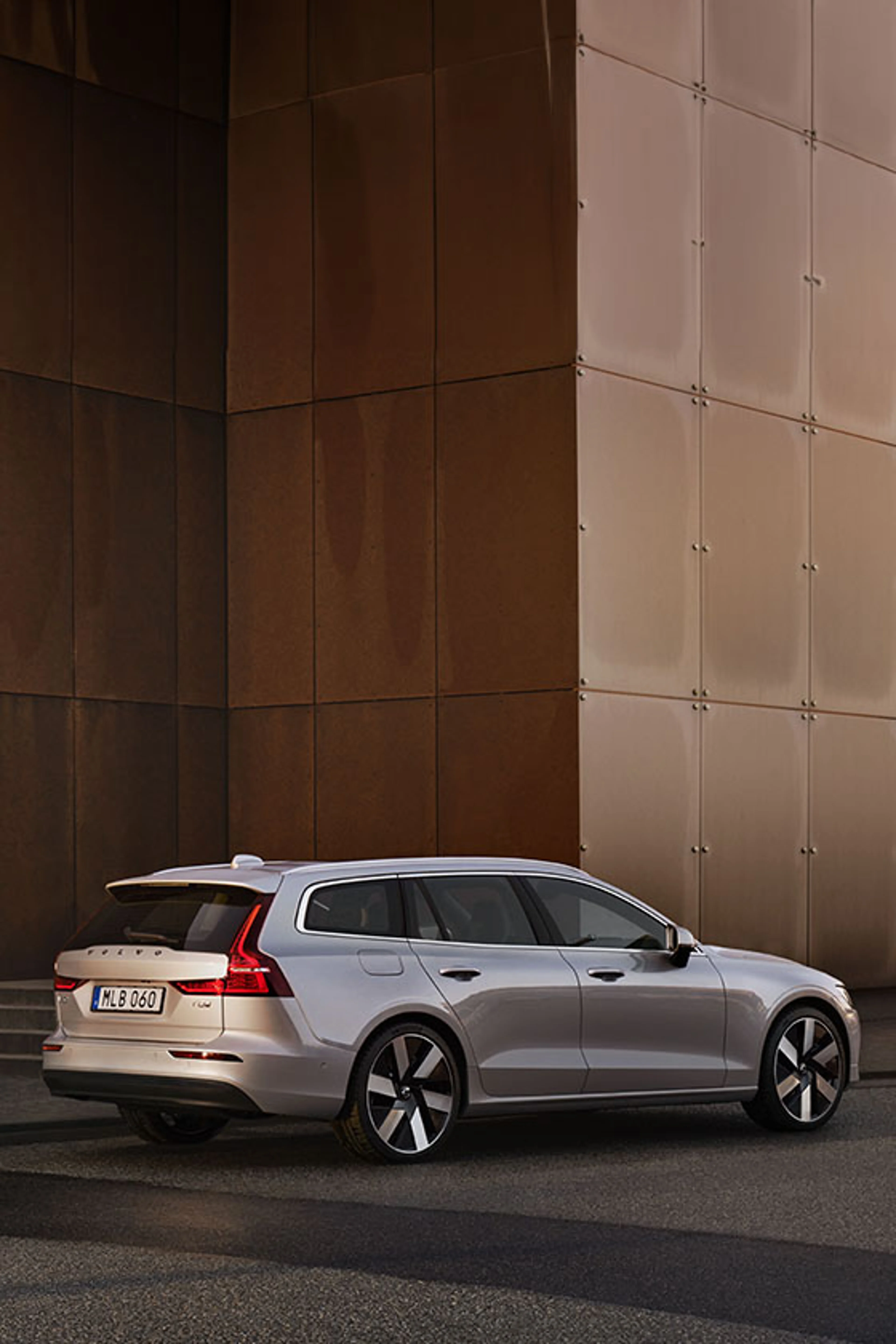 A silver Volvo V60 wagon is parked beside a modern, metallic building facade, viewed from the rear three-quarter angle in soft, evening light. The car's taillights are illuminated.