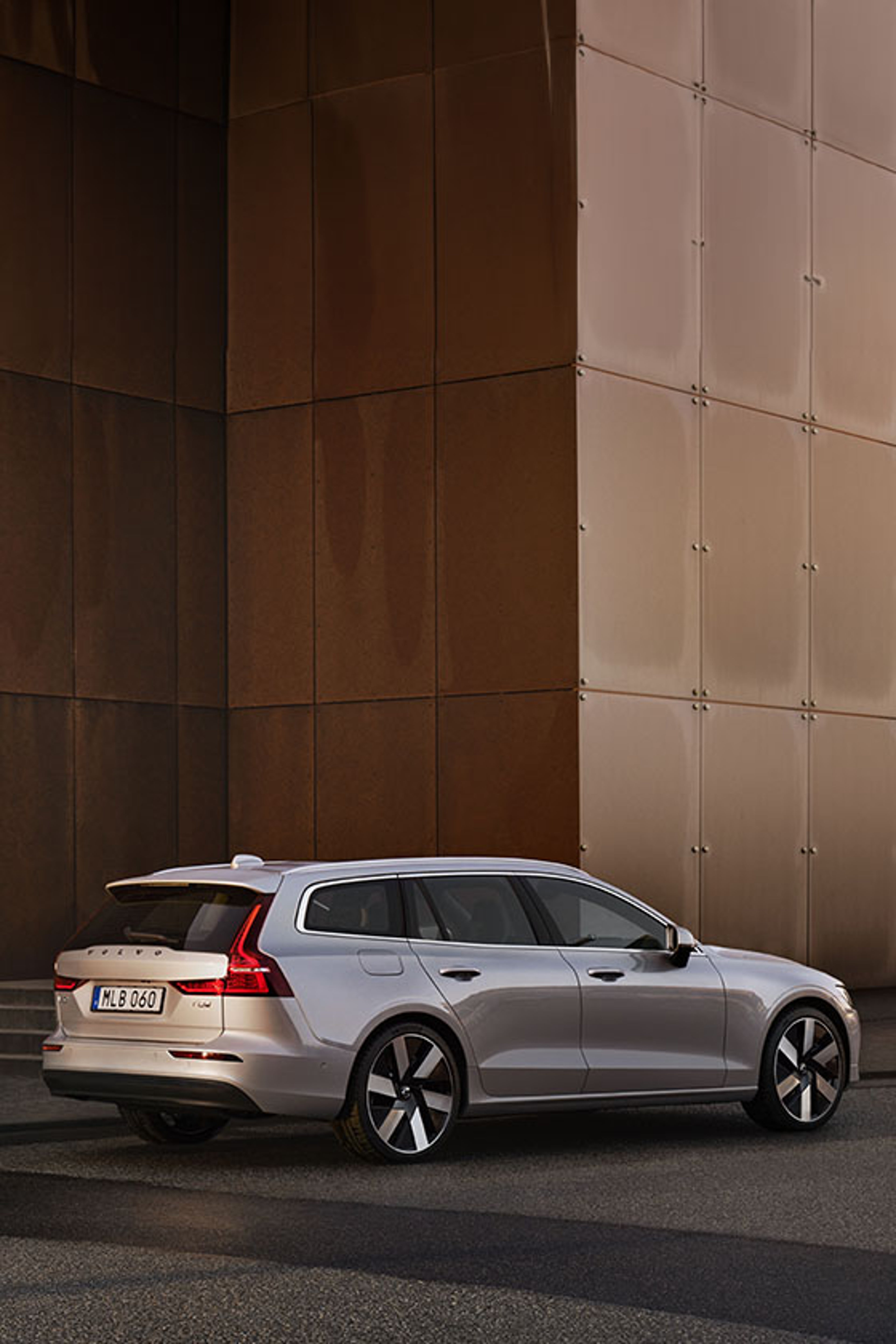 A silver Volvo V60 wagon is parked beside a modern building with brown and beige panels, showcasing its rear and side profile with distinctive taillights and alloy wheels.
