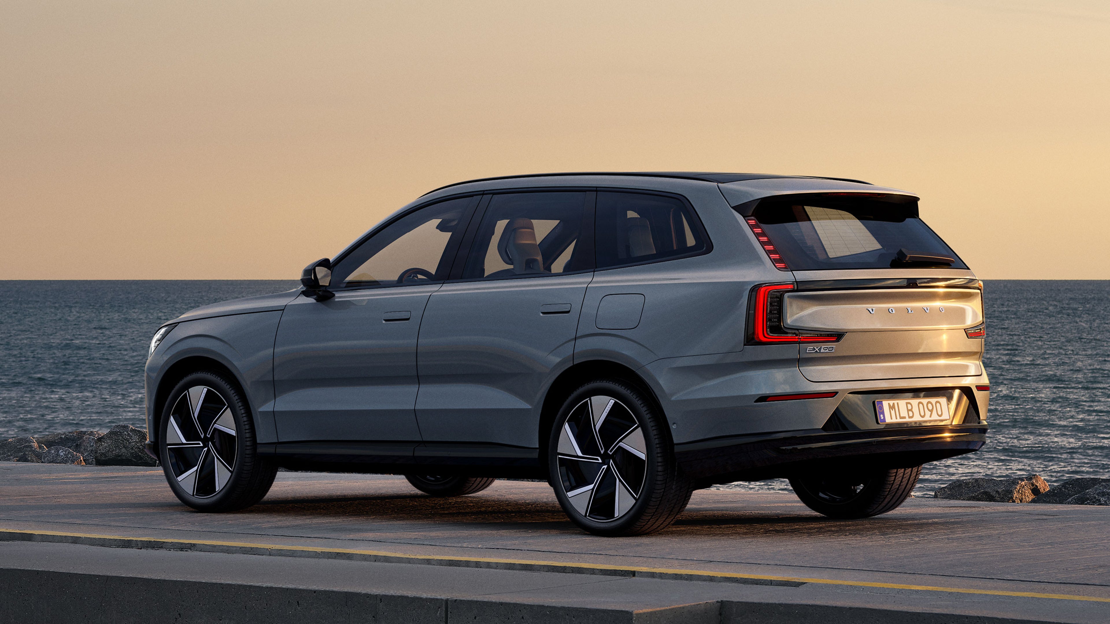 Vapour grey -värinen Volvo EX90 täyssähköinen katumaasturi pysäköitynä rantatien reunaan auringonlaskussa. Taustalla meri.