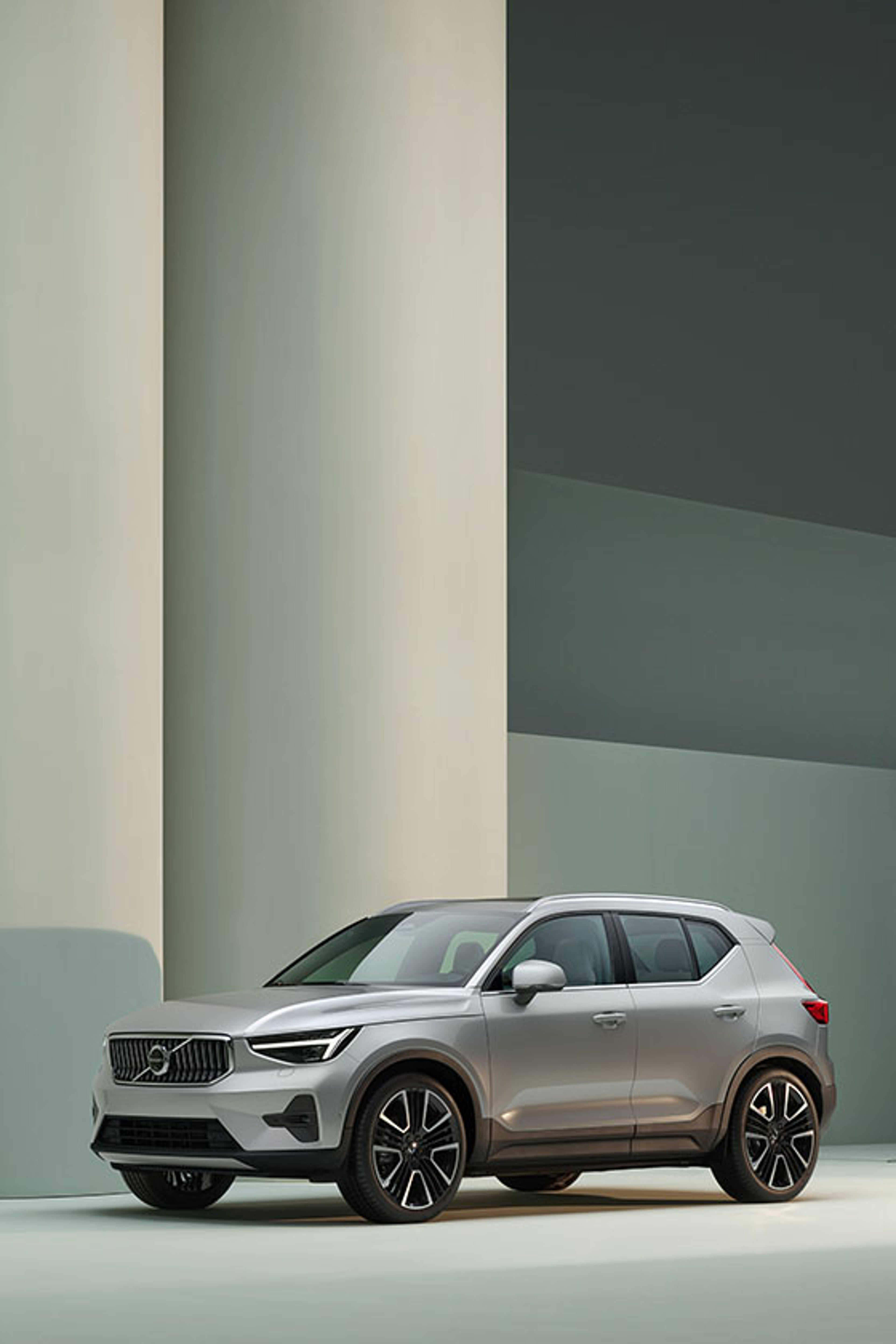 A silver Volvo XC40 SUV is parked in a minimalist, modern urban setting with large curved walls and a streetlamp, highlighting the vehicle’s sleek design and distinctive front grille.