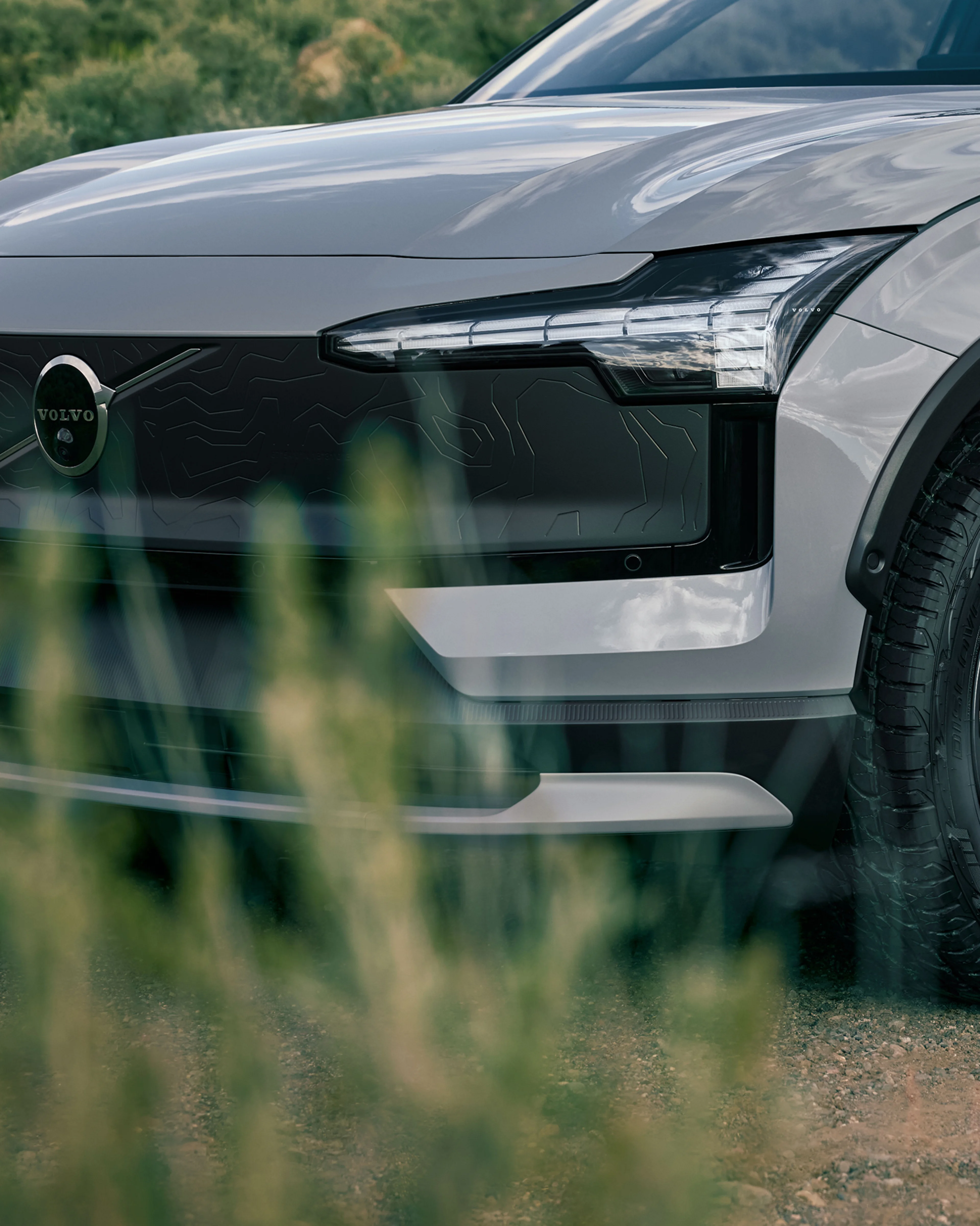 Κοντινή λήψη των τροχών παντός εδάφους και του εμπρός προφυλακτήρα του EX30 Cross Country πίσω από γρασίδι.