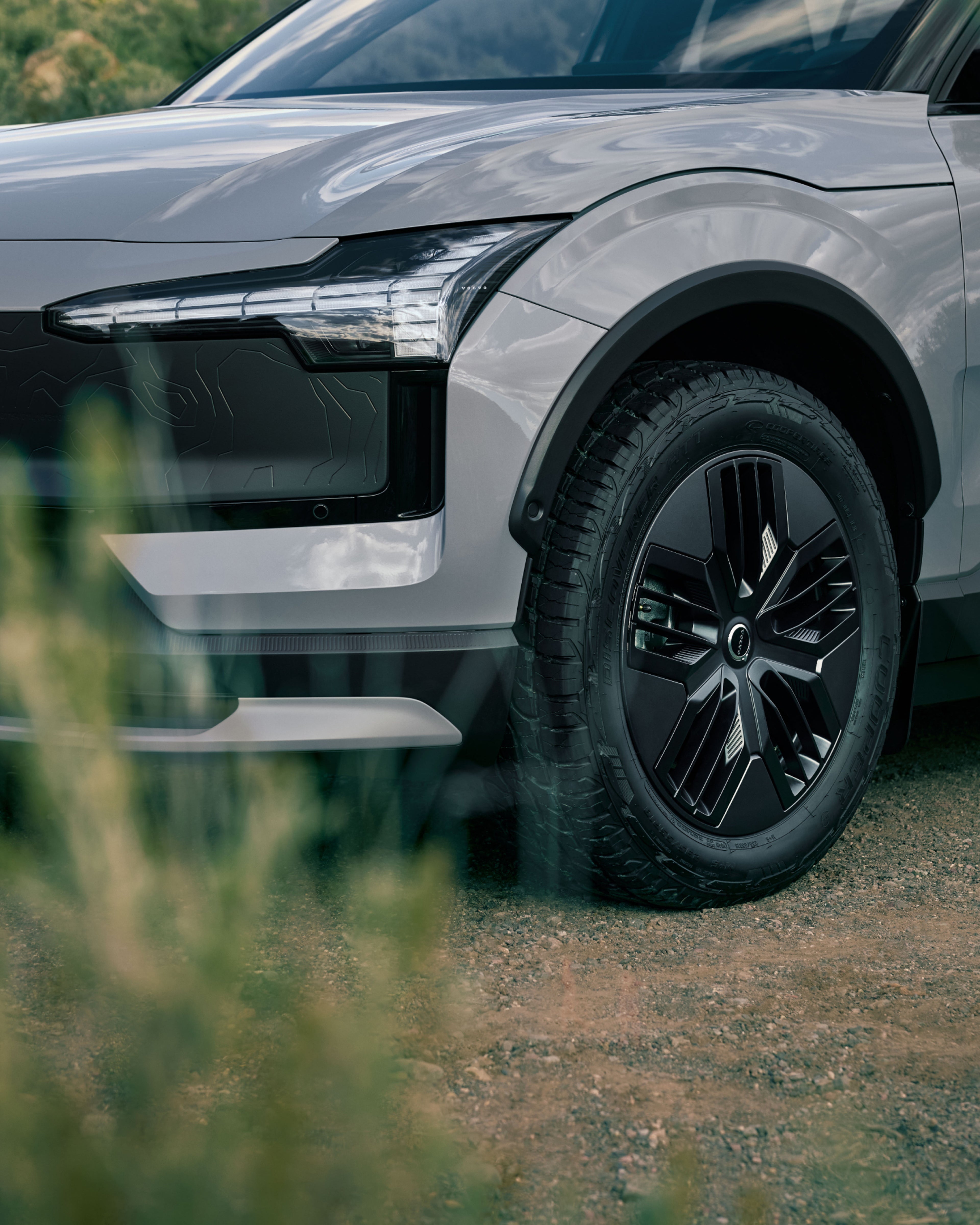 Close-up of the all-terrain wheels and front bumper on the EX30 Cross Country behind some grass.