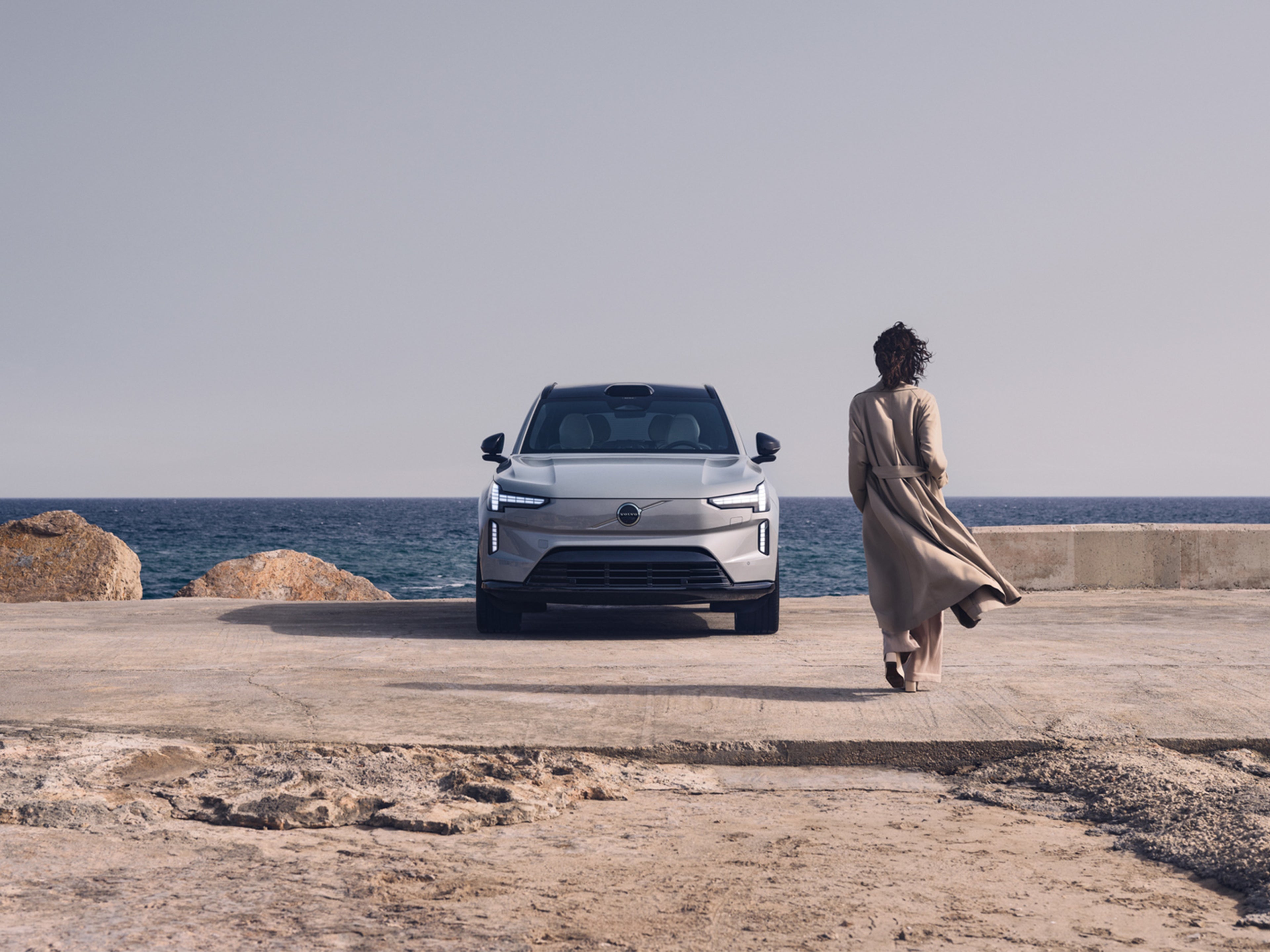 Trenssiin pukeutunut nainen kävelee kohti merenrantaan pysäköityä Volvo EX90 täyssähköautoa.