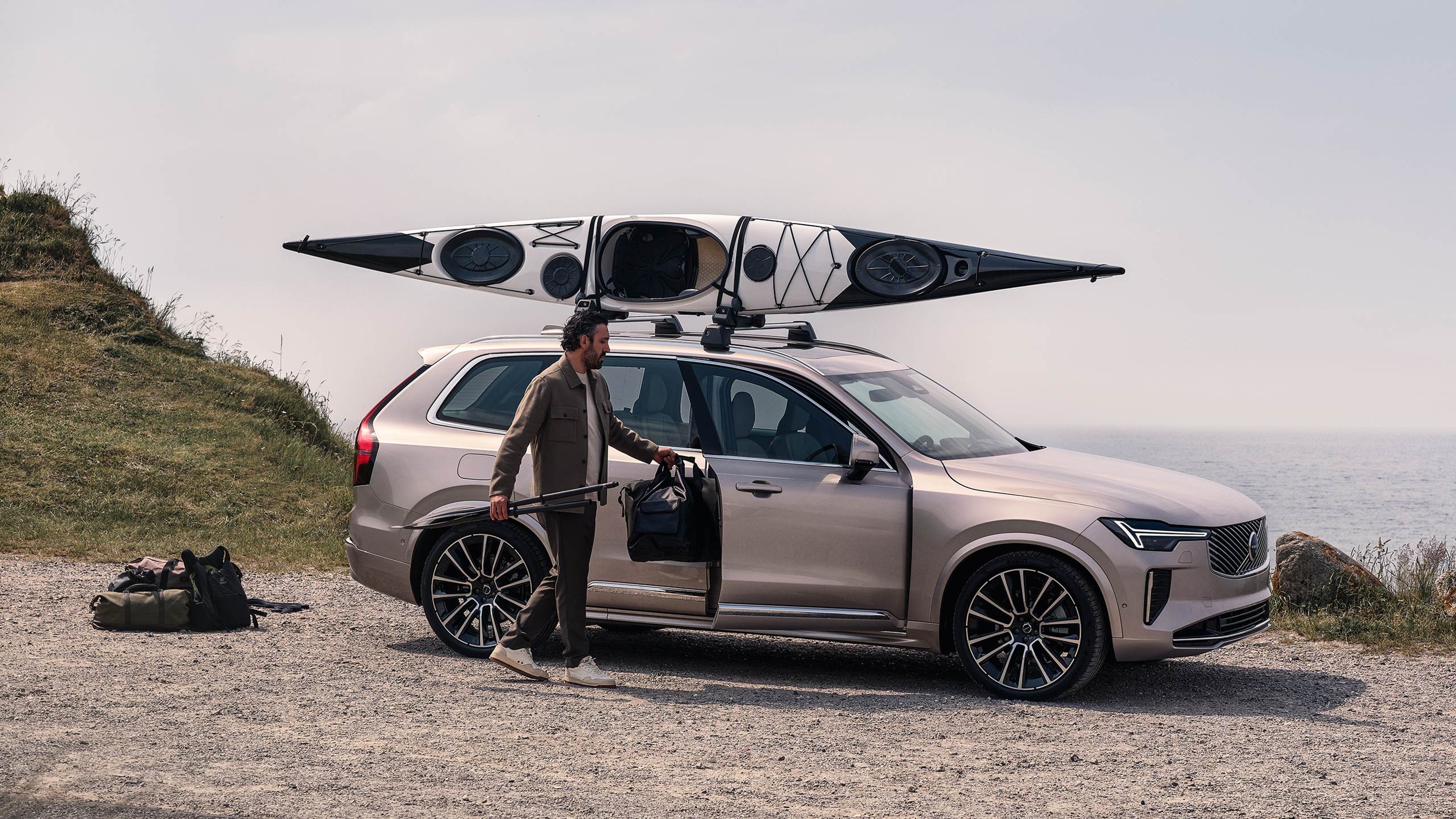 Mies pakkaa rantatien varteen pysäköityä Bright dusk -väristä Volvo XC90 lataushybridikatumaasturia, jonka taakkatelineisiin on kiinnitetty kajakki.