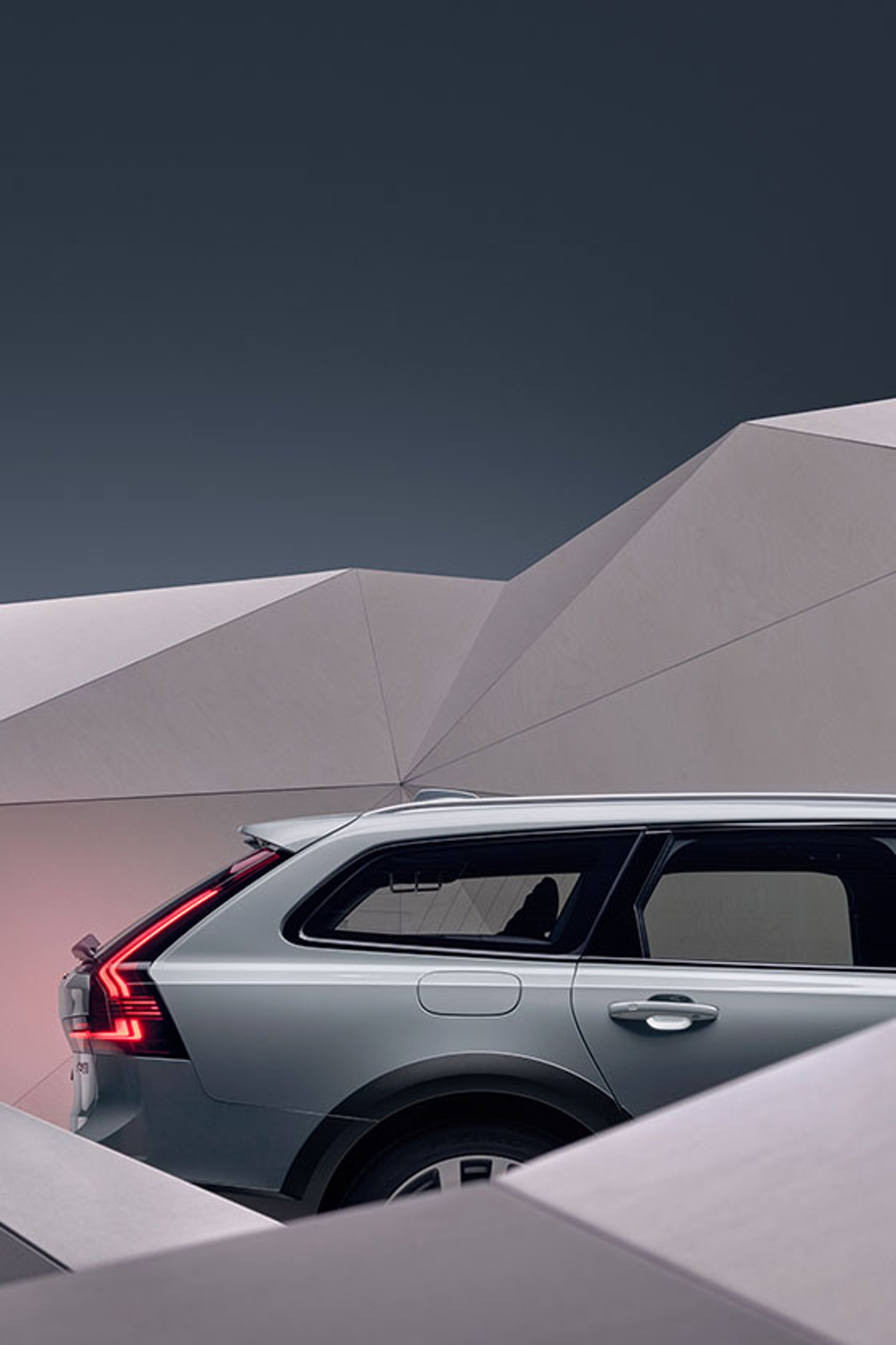 A silver Volvo V90 station wagon is partially visible, framed by angular, geometric structures. The image highlights the car’s rear side, distinctive taillight, and roofline design.