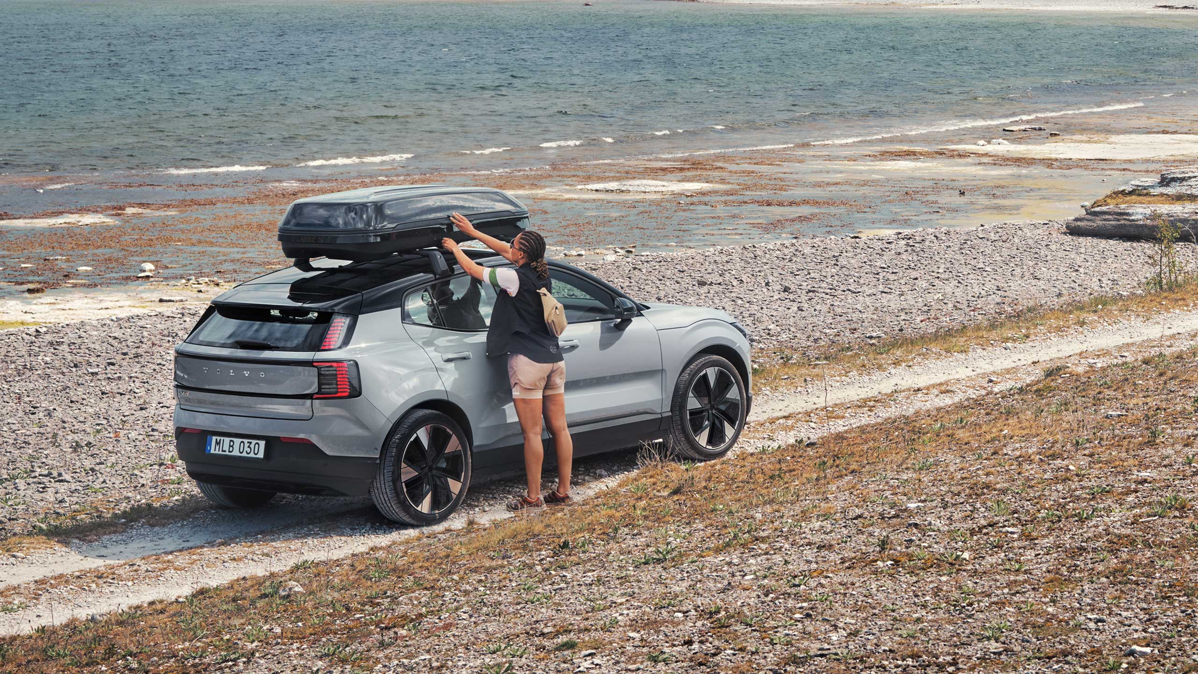 Nainen avaa rantatien varteen pysäköidyn Cloud blue -värisen Volvo EX30 täyssähköisen katumaasturin taakkatelineisiin kiinnitettyä kattolaatikkoa.