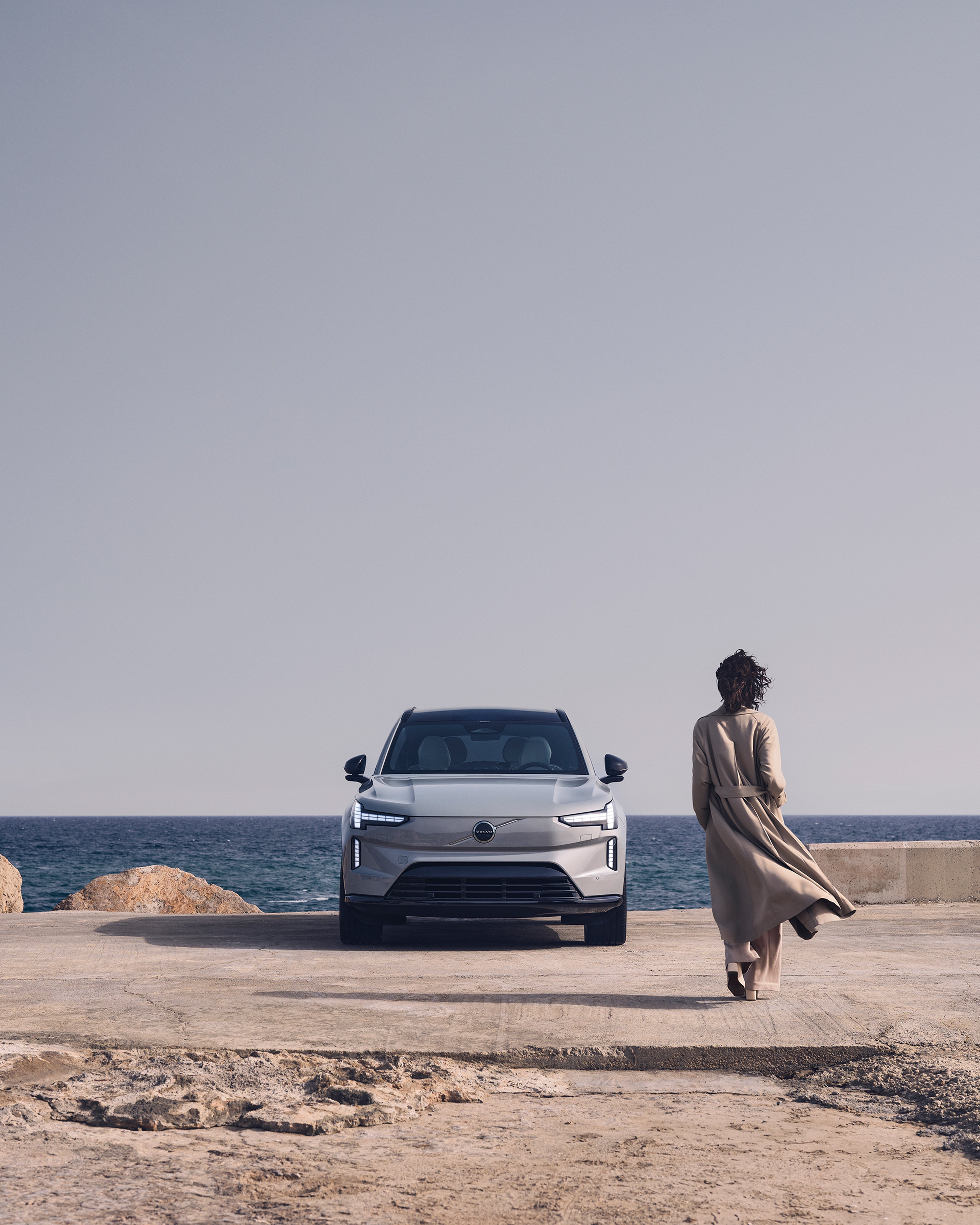 Eine Frau geht auf einen geparkten EX90 zu, der am Rande einer Küstenstraße steht.