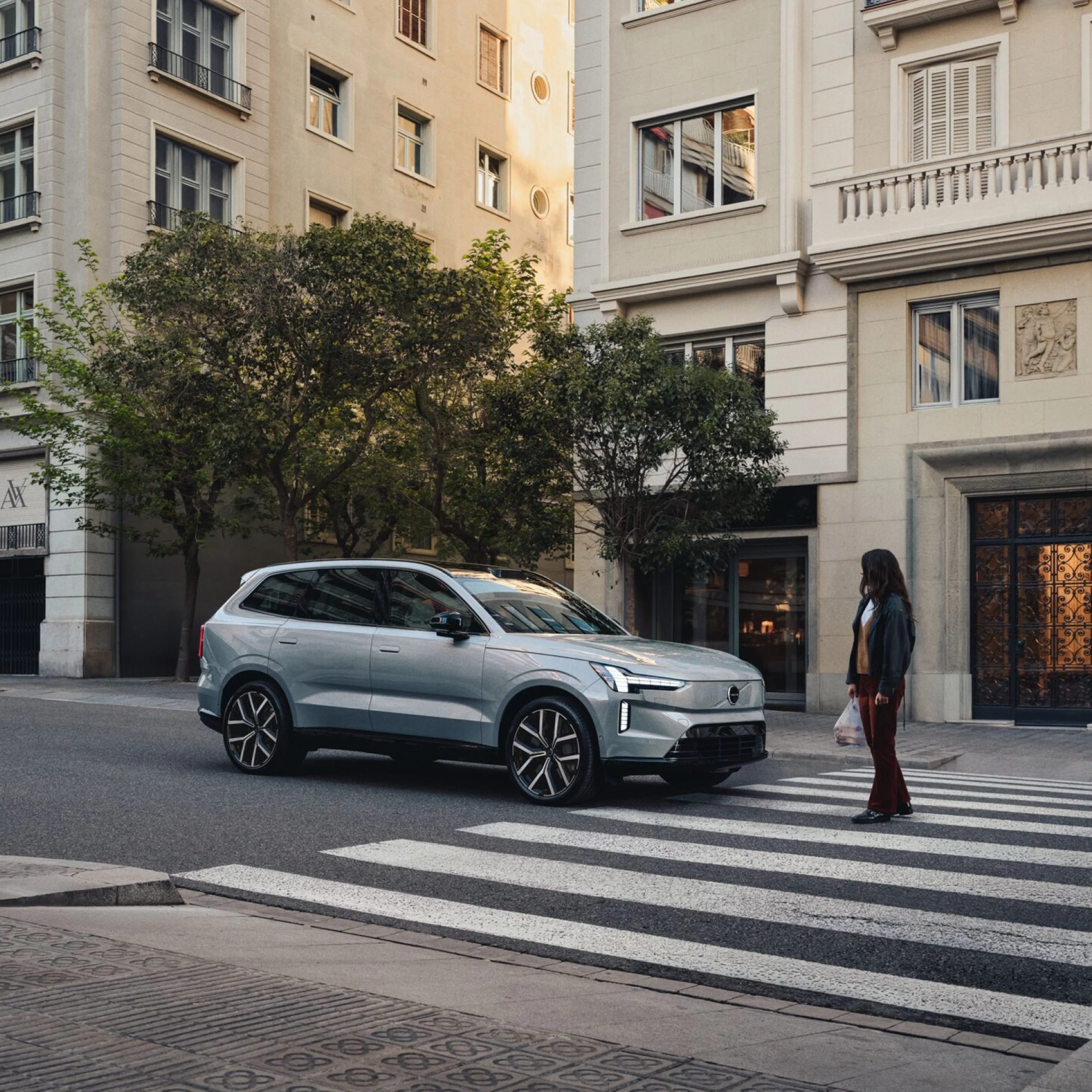 Une femme traverse sur le passage piéton dans un environnement urbain. Une Volvo EX90 gris clair s'arrête devant le passage piéton.