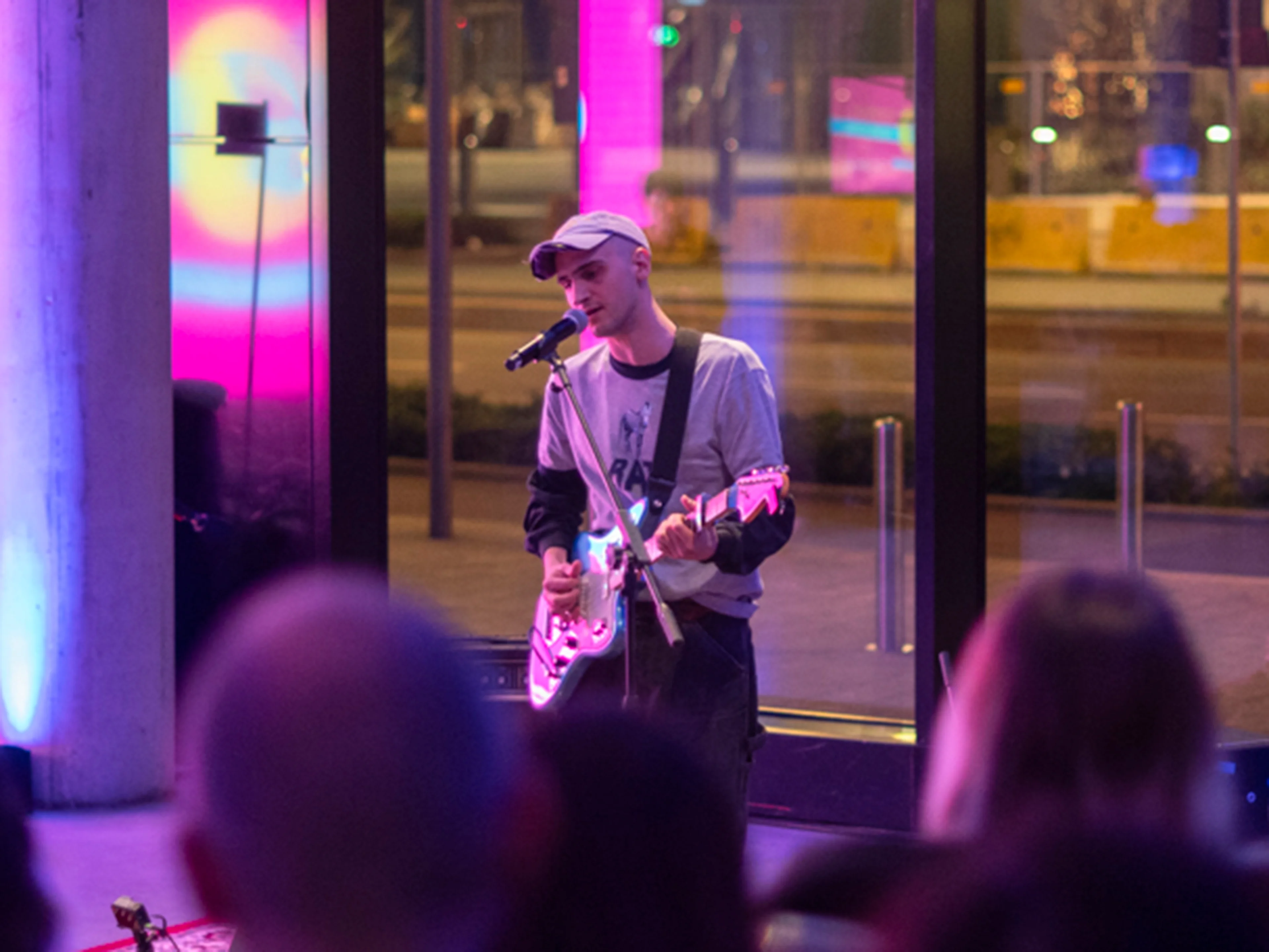 A musician plays an electric guitar and sings indoors, illuminated by colorful purple and blue lights, with an audience watching and large glass windows showing a street outside.