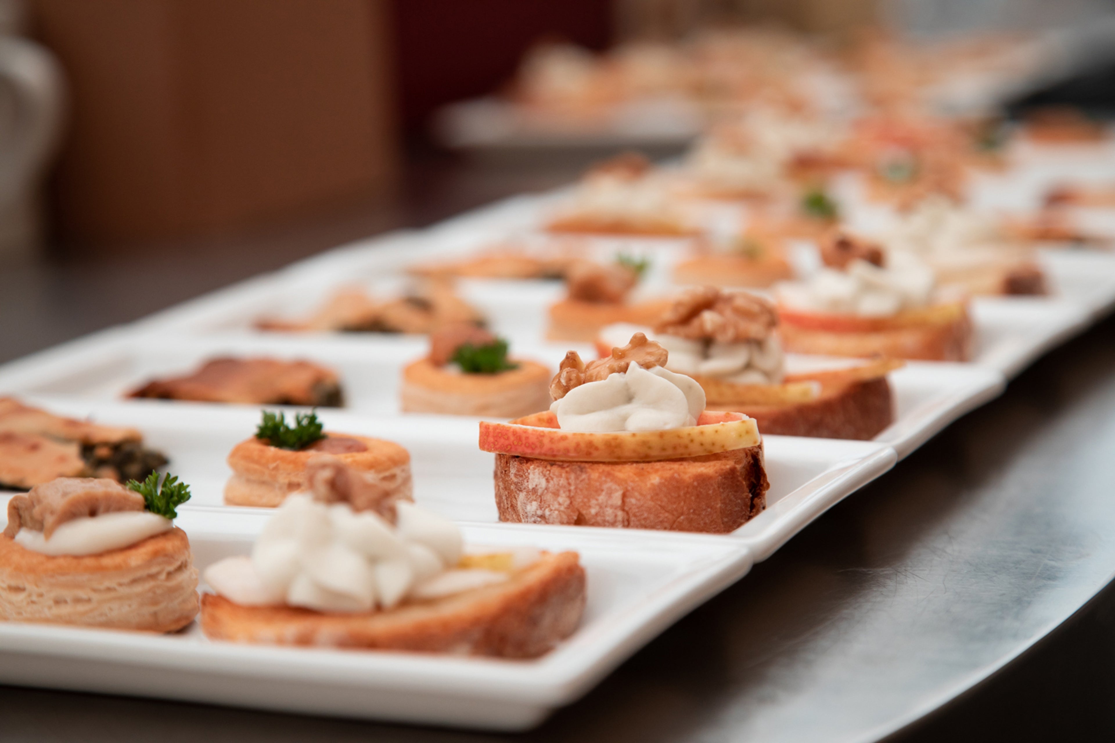 Close-up of assorted appetizers arranged on a white rectangular platter, featuring bread slices topped with cream, apple, and walnuts, and other bite-sized savory pastries garnished with herbs.
