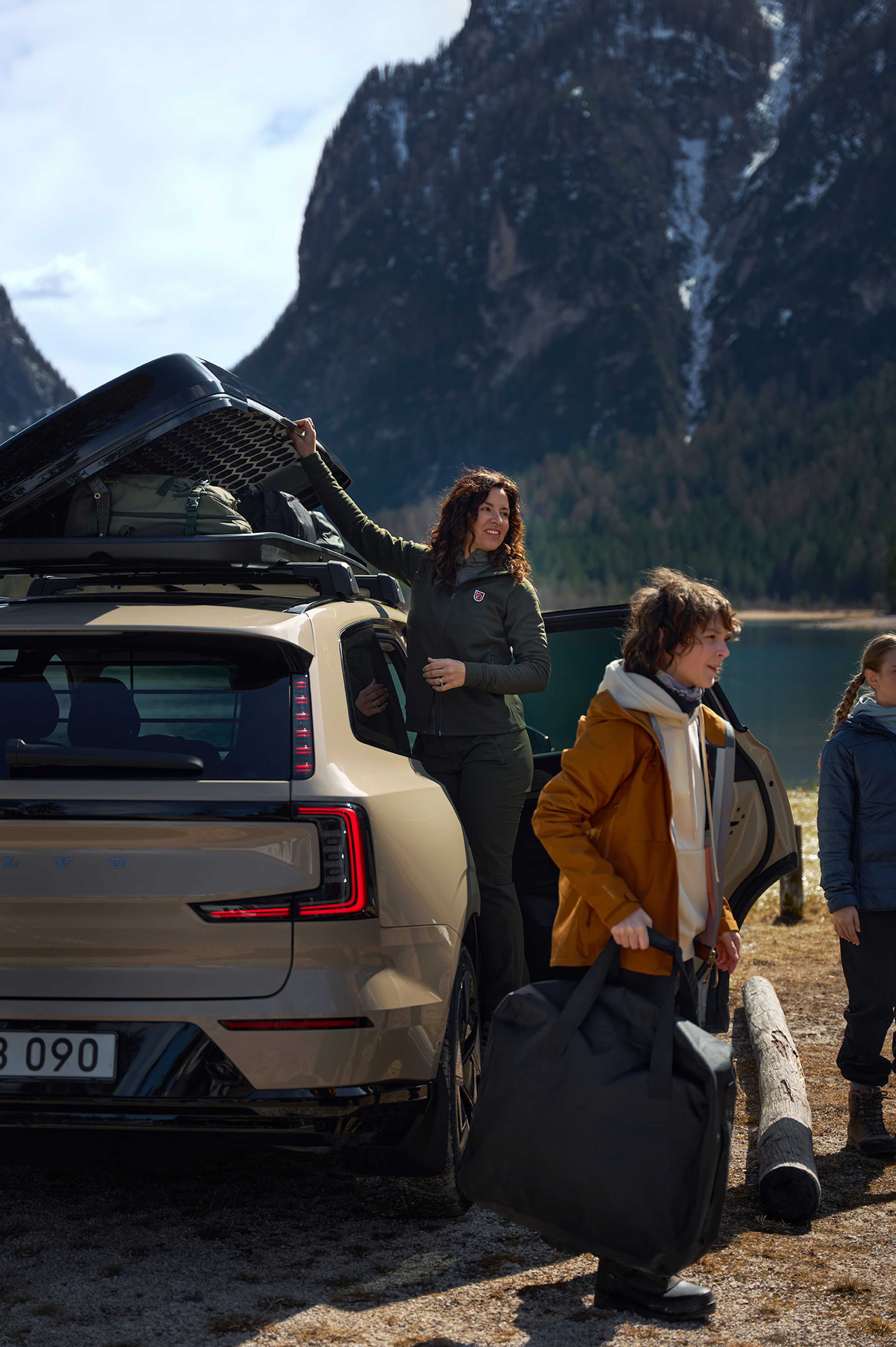 A woman holds open a filled cargo roof box on a Volvo SUV, with two children looking towards nature.
