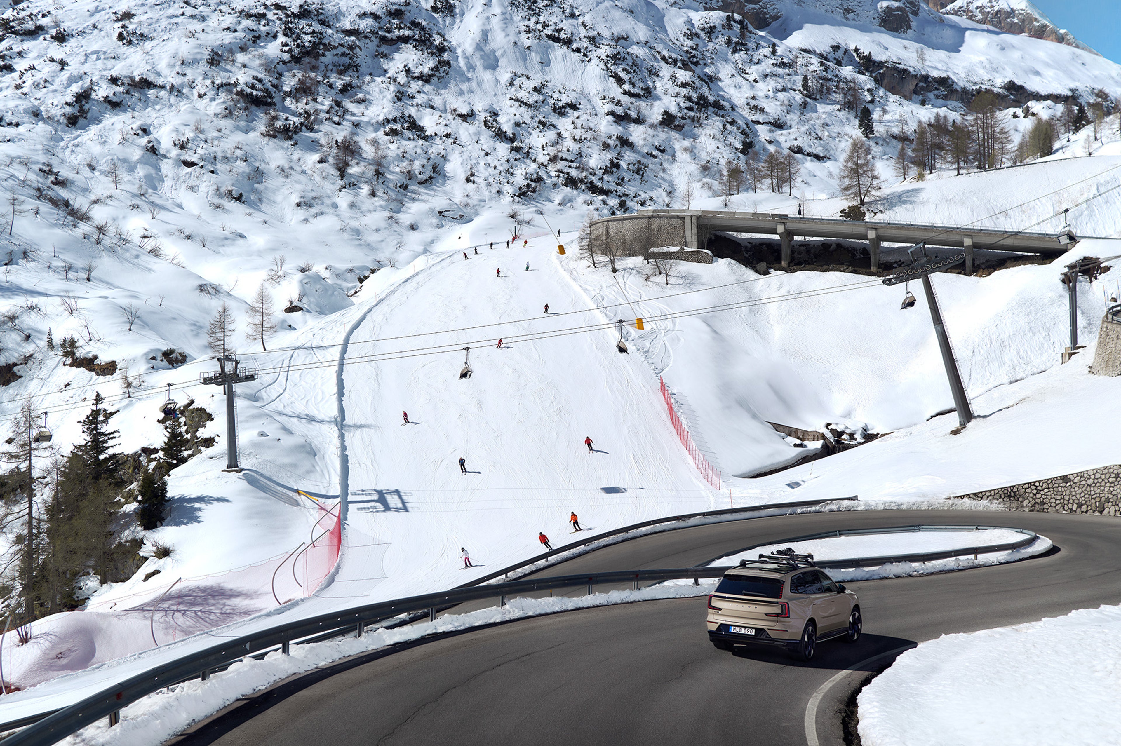 A Volvo EX90 travels down a winding road in winter with a ski slope in the background.
