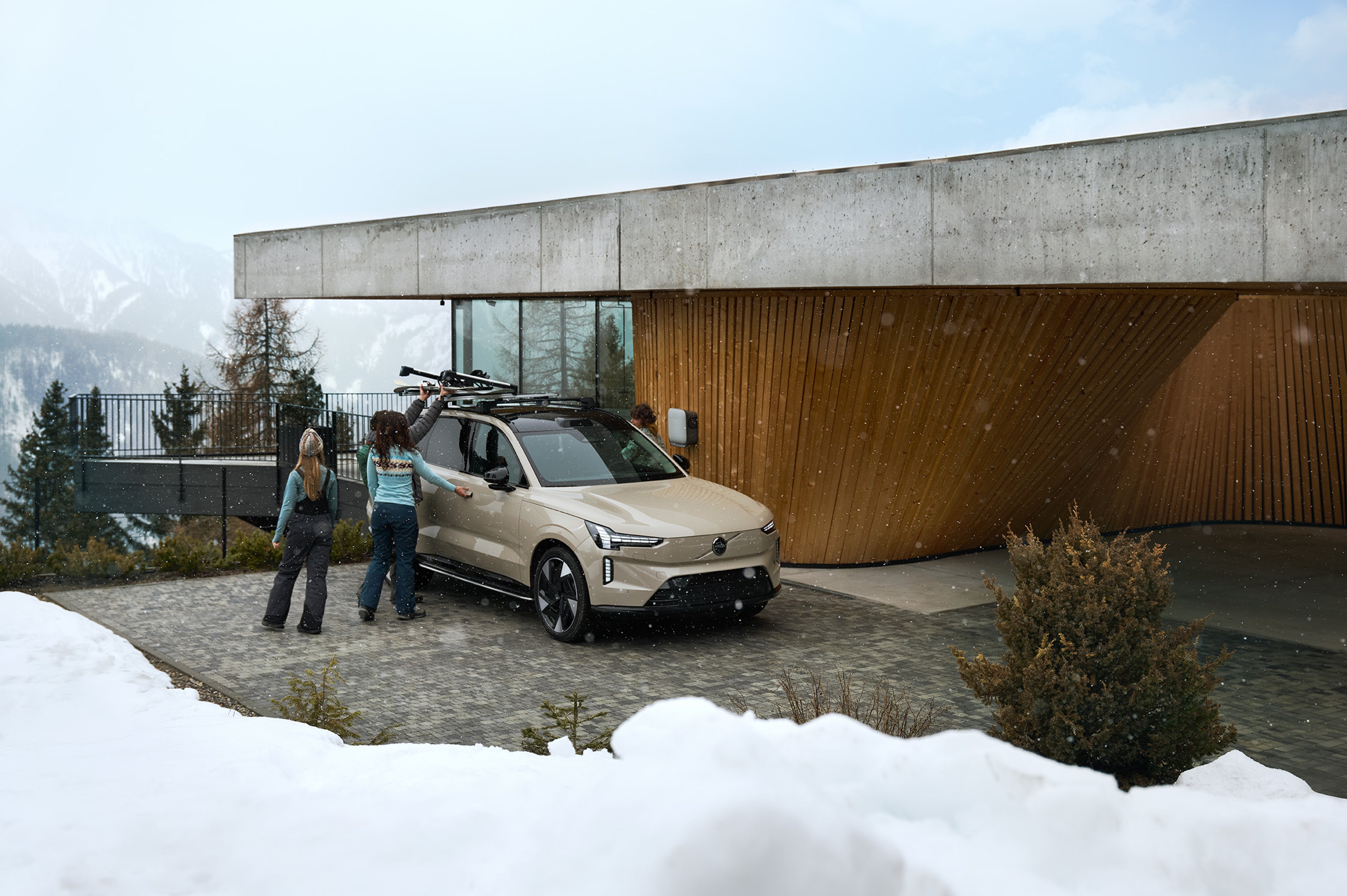 Ryhmä ihmisiä lastaamassa suksia Volvo EX90:n sisäänvedettävään suksipidikkeeseen, auto pysäköitynä talon eteen talvisessa maisemassa.