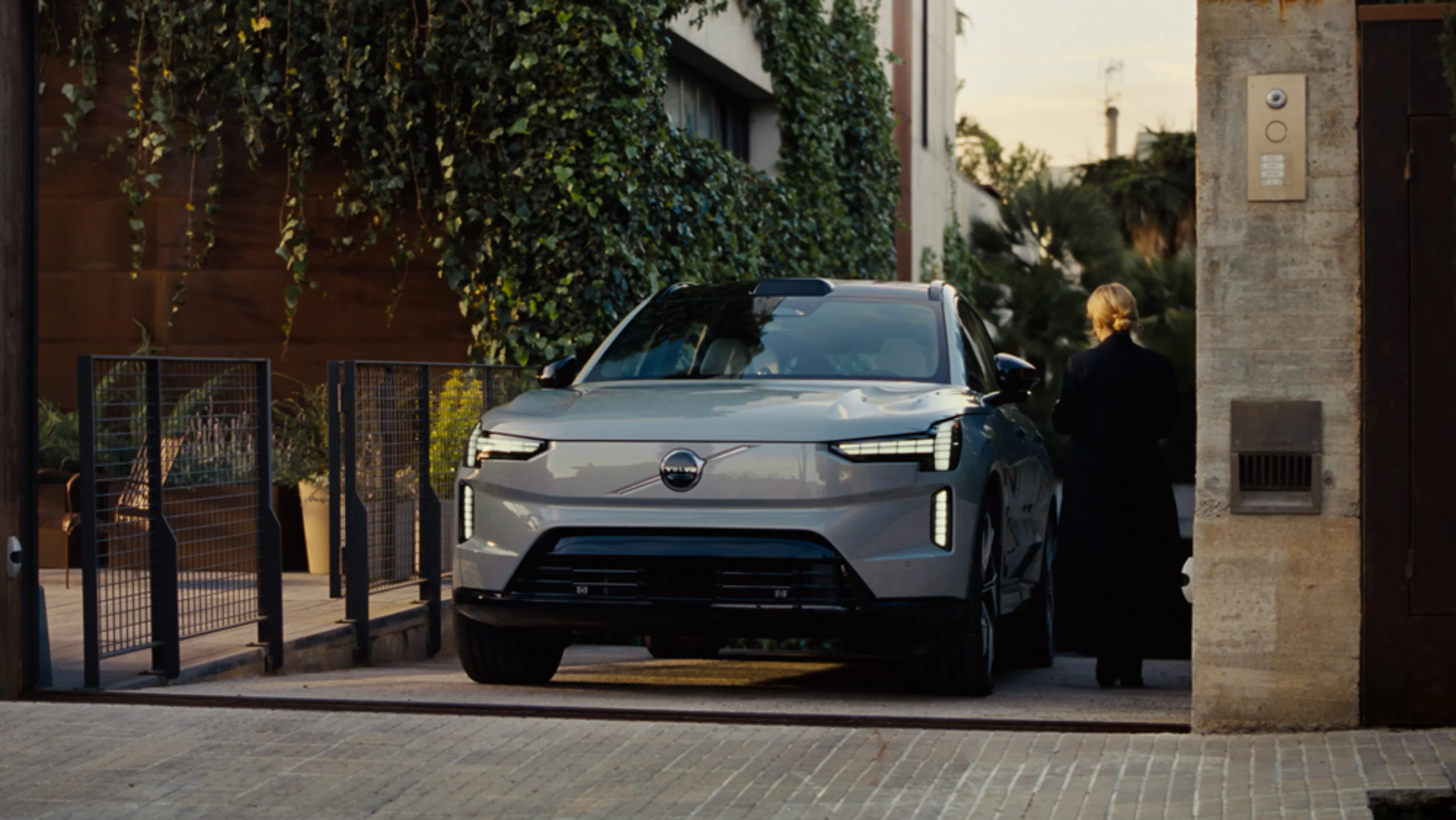 A white Volvo EX30 compact SUV is parked on the street beside a light-colored wall, viewed from the side. The car features black accents and distinctive alloy wheels.