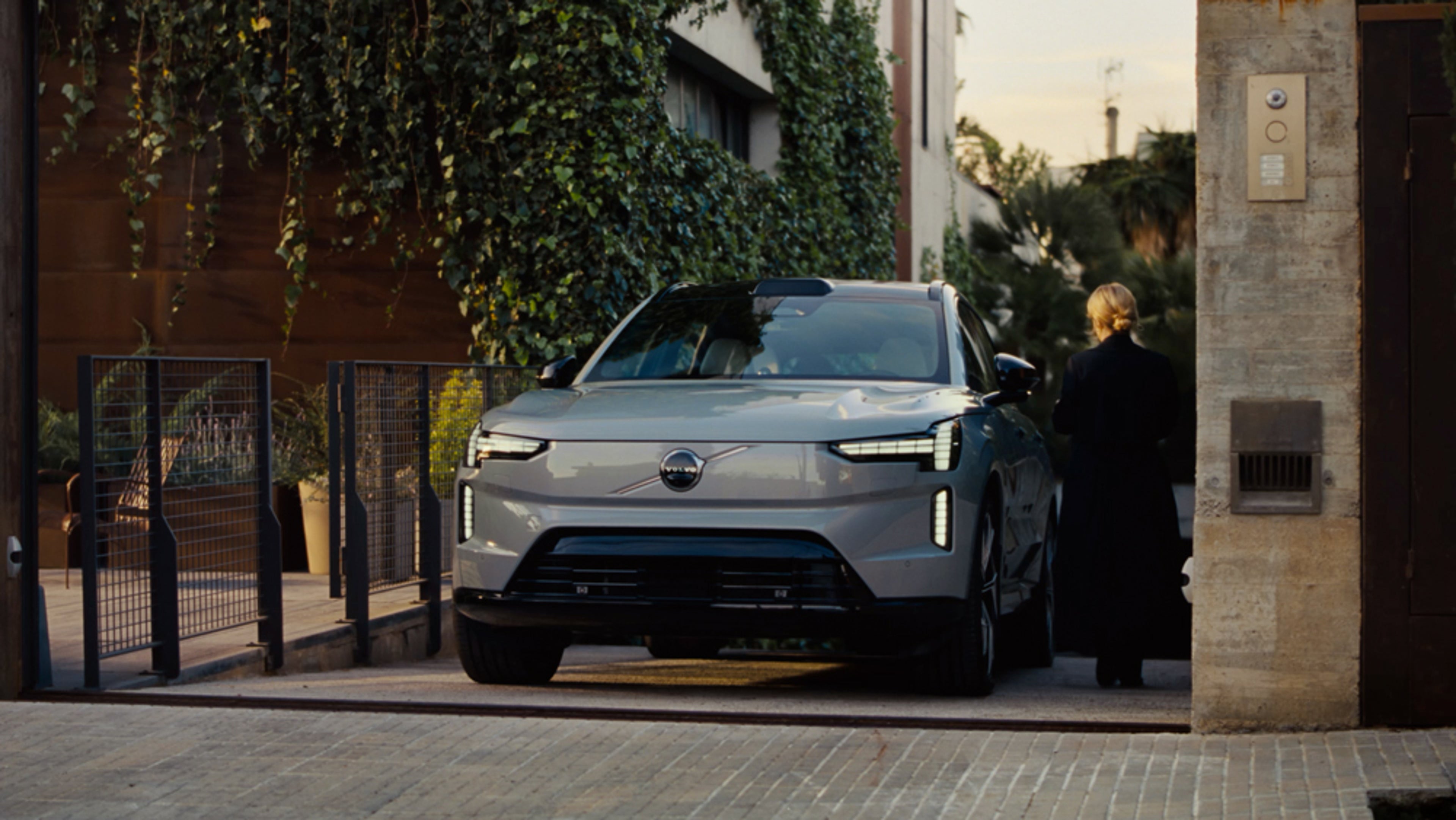 A white Volvo EX30 compact SUV is parked on the street beside a light-colored wall, viewed from the side. The car features black accents and distinctive alloy wheels.
