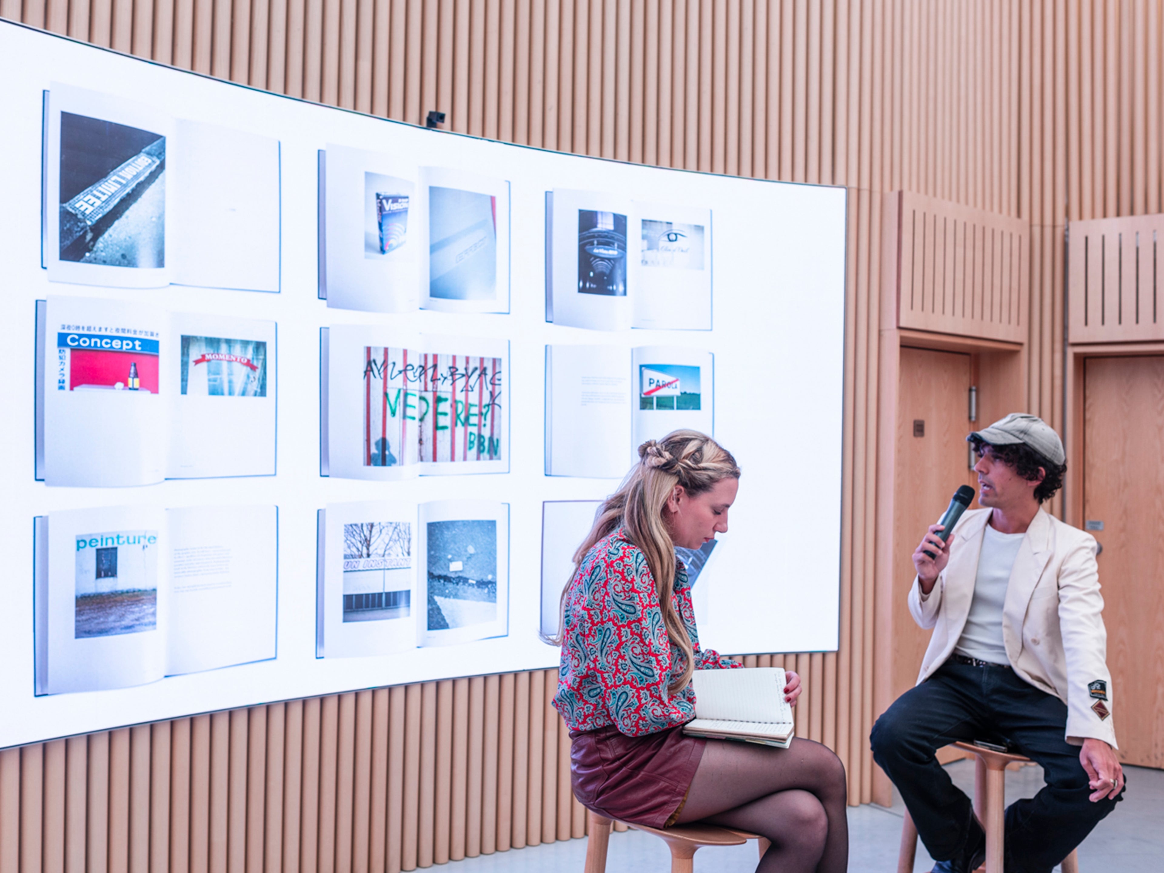 Two people sit on stools in front of a large screen displaying various book or magazine covers and photographs, inside a modern room with wooden paneling.