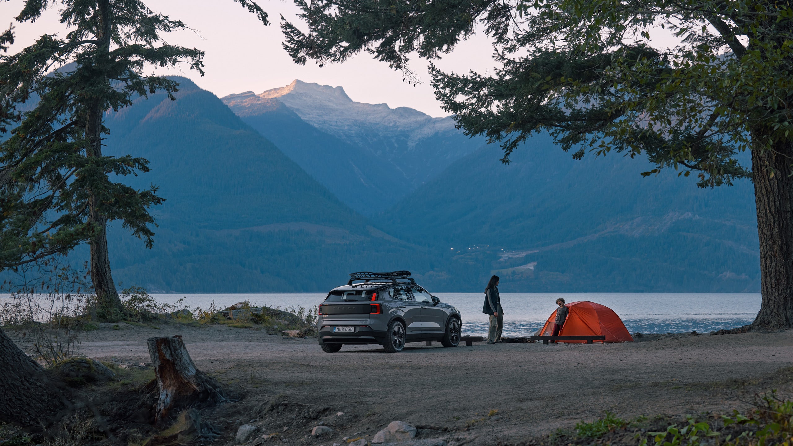 En campingplats med ett orange tält vid en sjö och höga berg i bakgrunden. En EX30 Cross Country, en kvinna och ett barn är vid tältet.