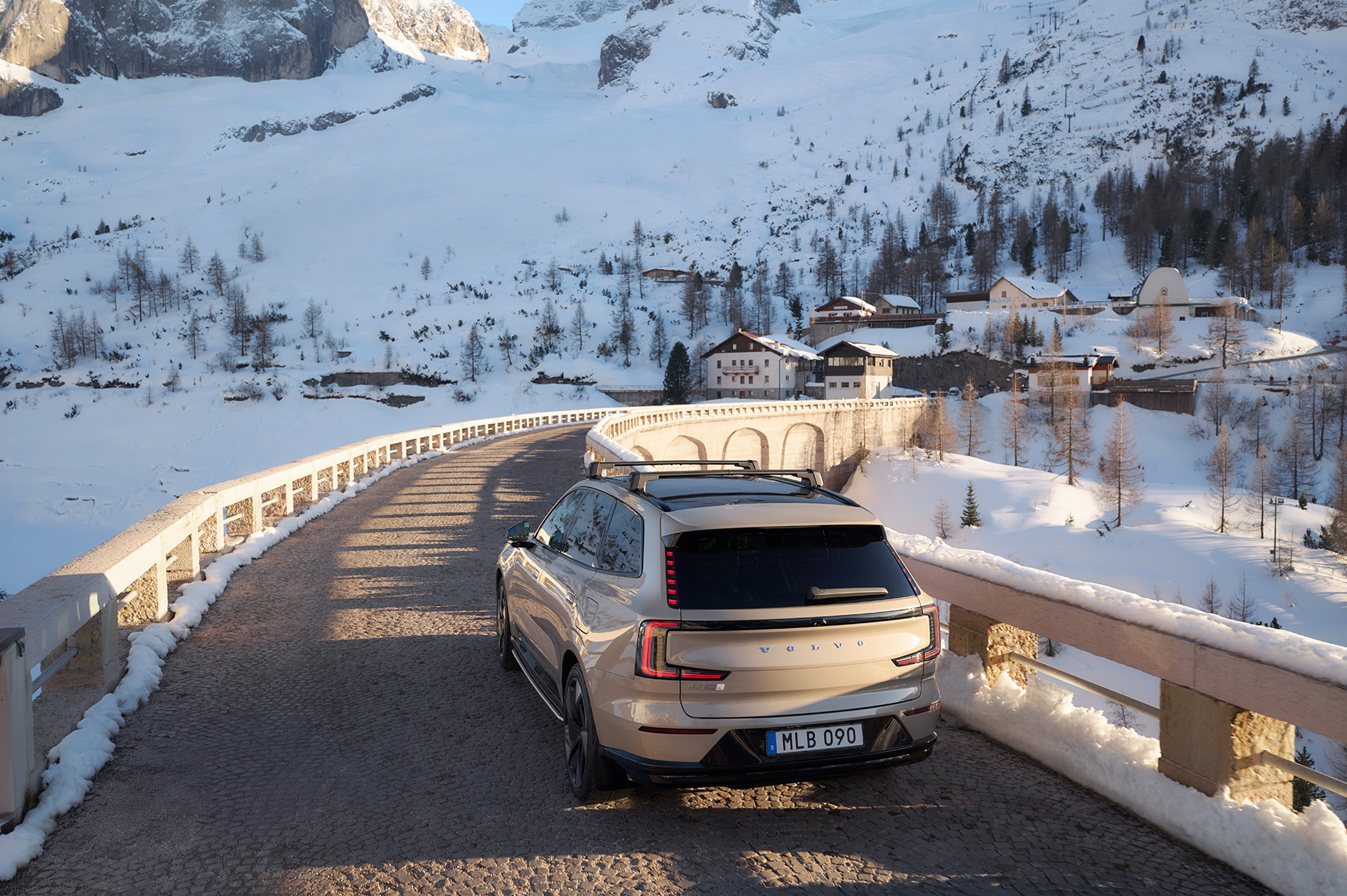 A Volvo EX90 driving over a bridge towards snow-covered buildings.