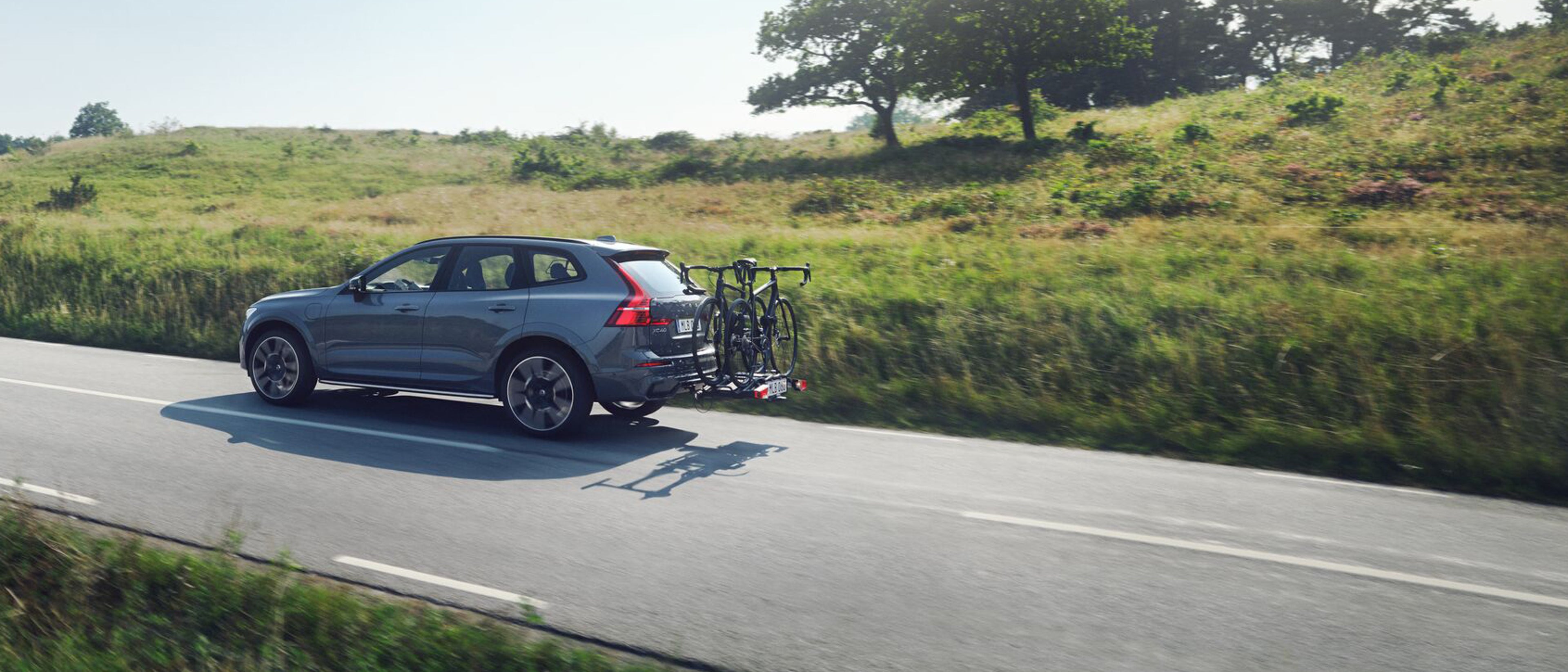 En sølvfarvet Volvo XC60 SUV kører ad en naturskøn, tom vej omgivet af frodig grøn beplantning og træer under en klar himmel.
