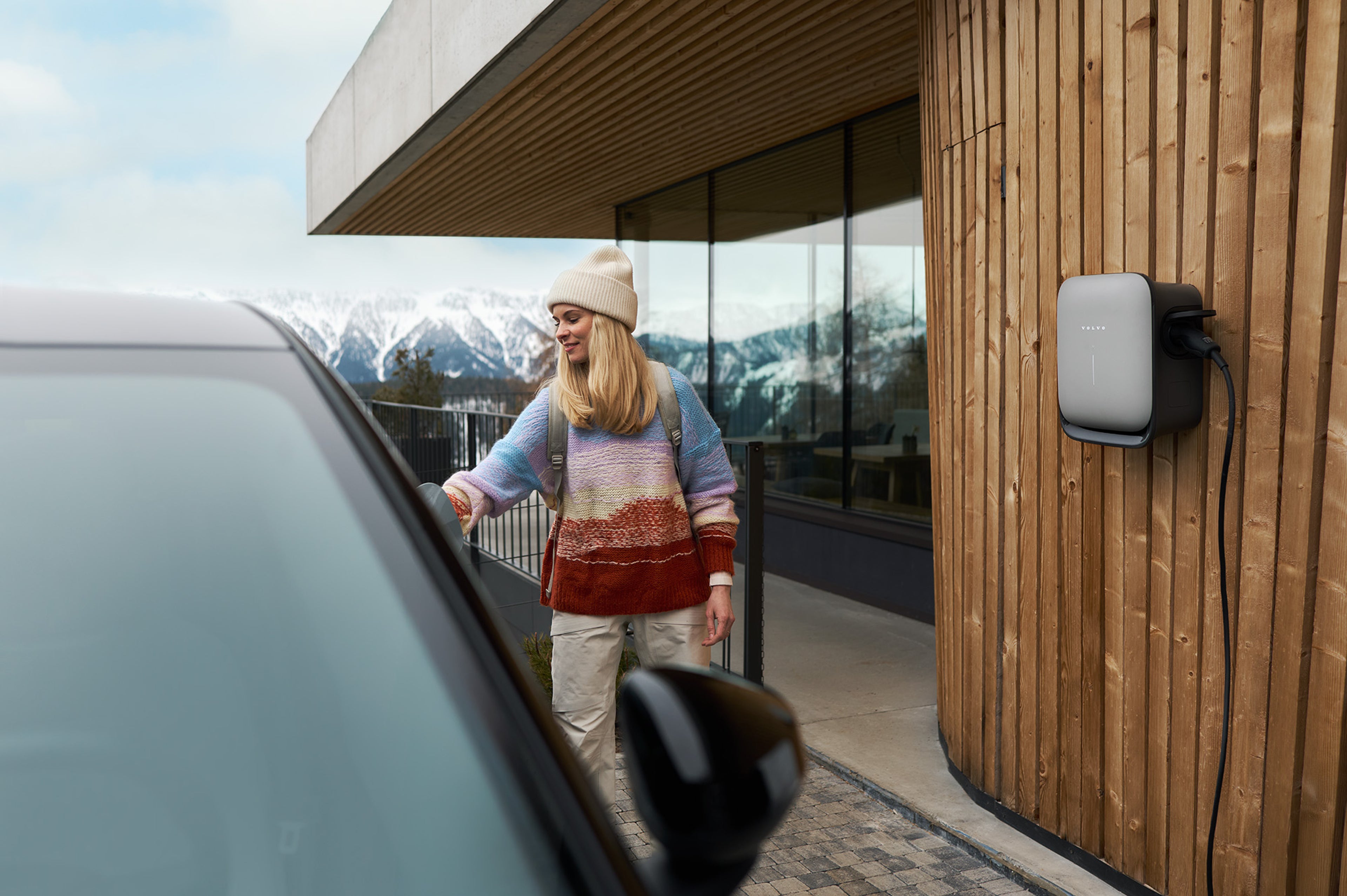 A woman uses a charging cable plugged into a Volvo Wallbox at a home with snowy mountains in background.