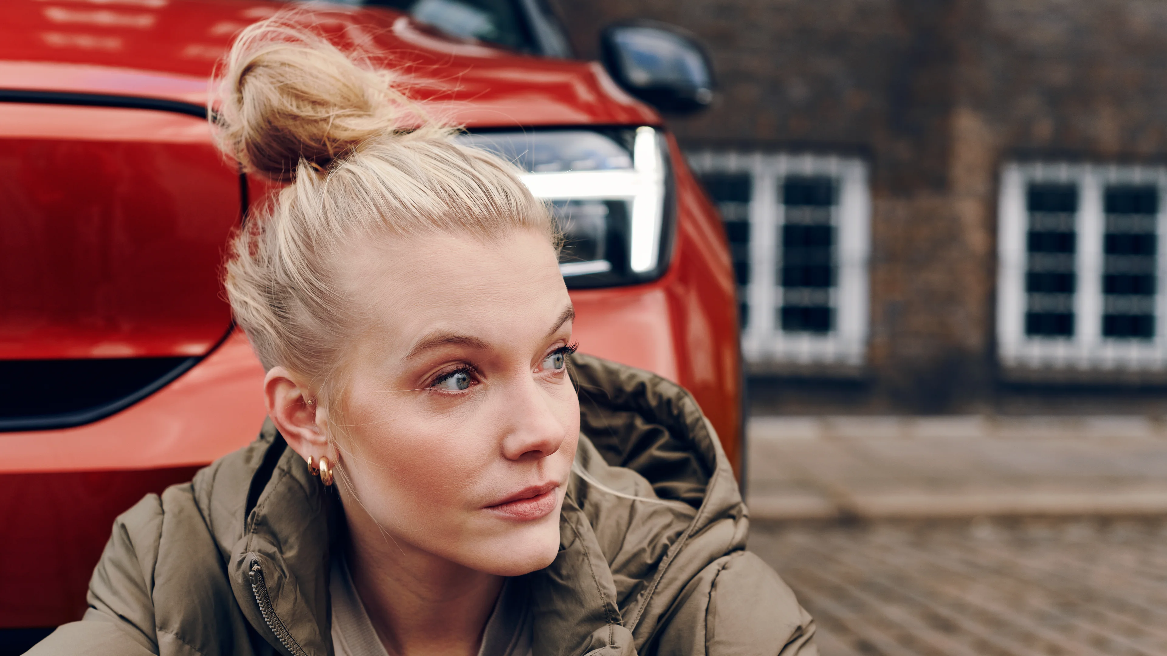A red Volvo XC40 is parked on a cobblestone street, with its front grille and headlight visible next to a person wearing a green jacket sitting nearby.