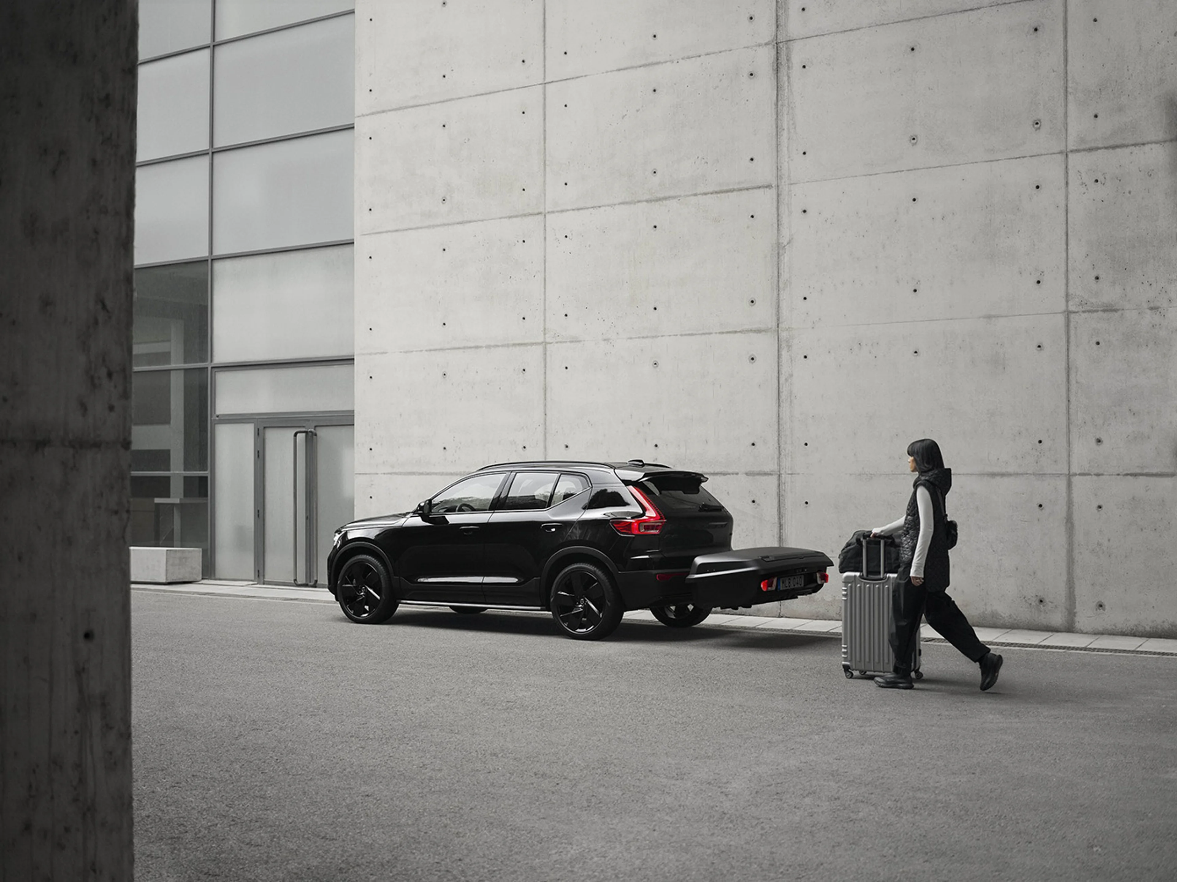 Eine Frau mit einem Koffer geht auf die Ablagebox für die Anhängerkupplung ihres Volvo zu.