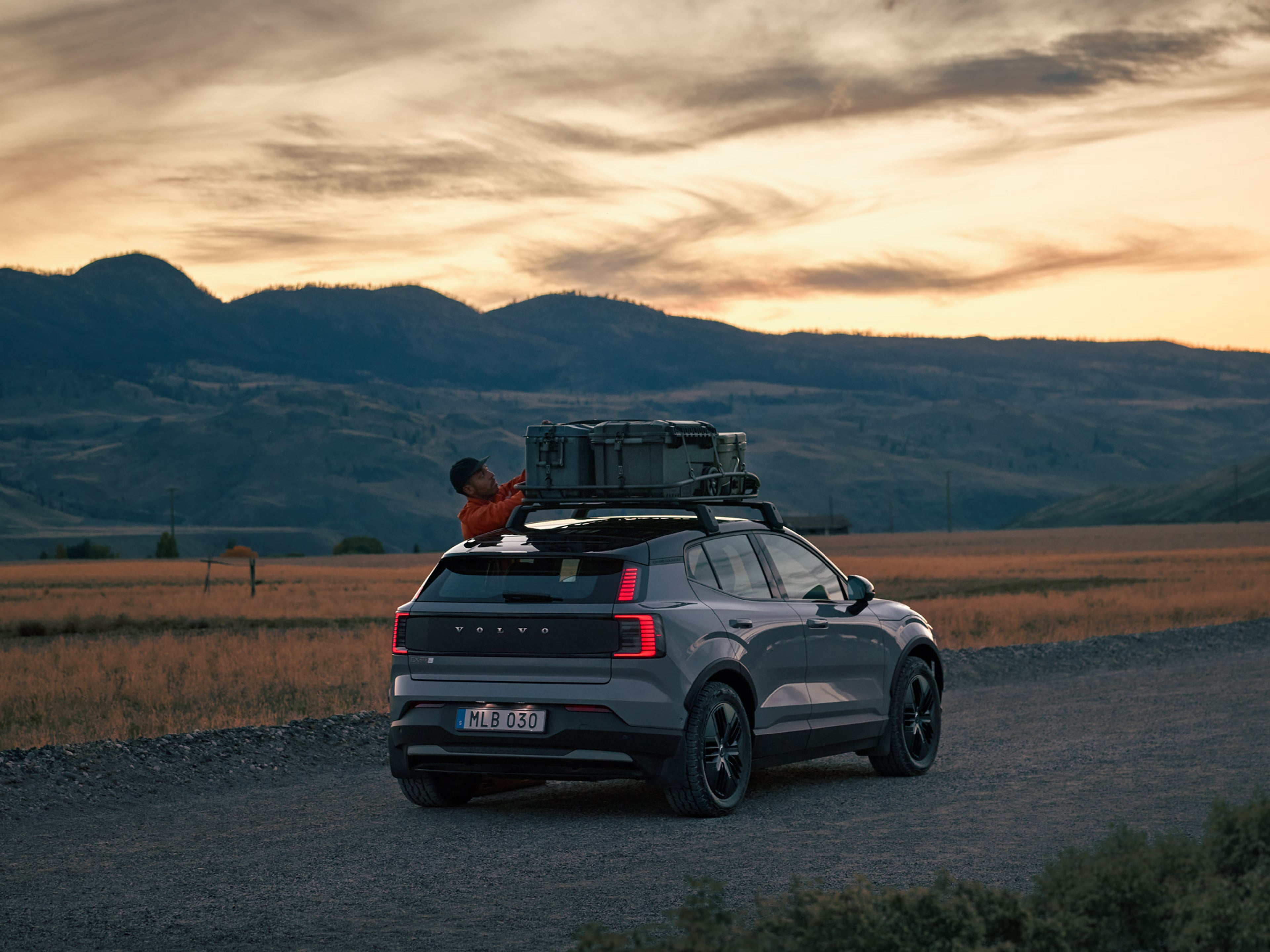 En person lastar en takkorg på en Volvo EX30 Cross Country fotograferad snett bakifrån på en grusväg i skymningsljus.