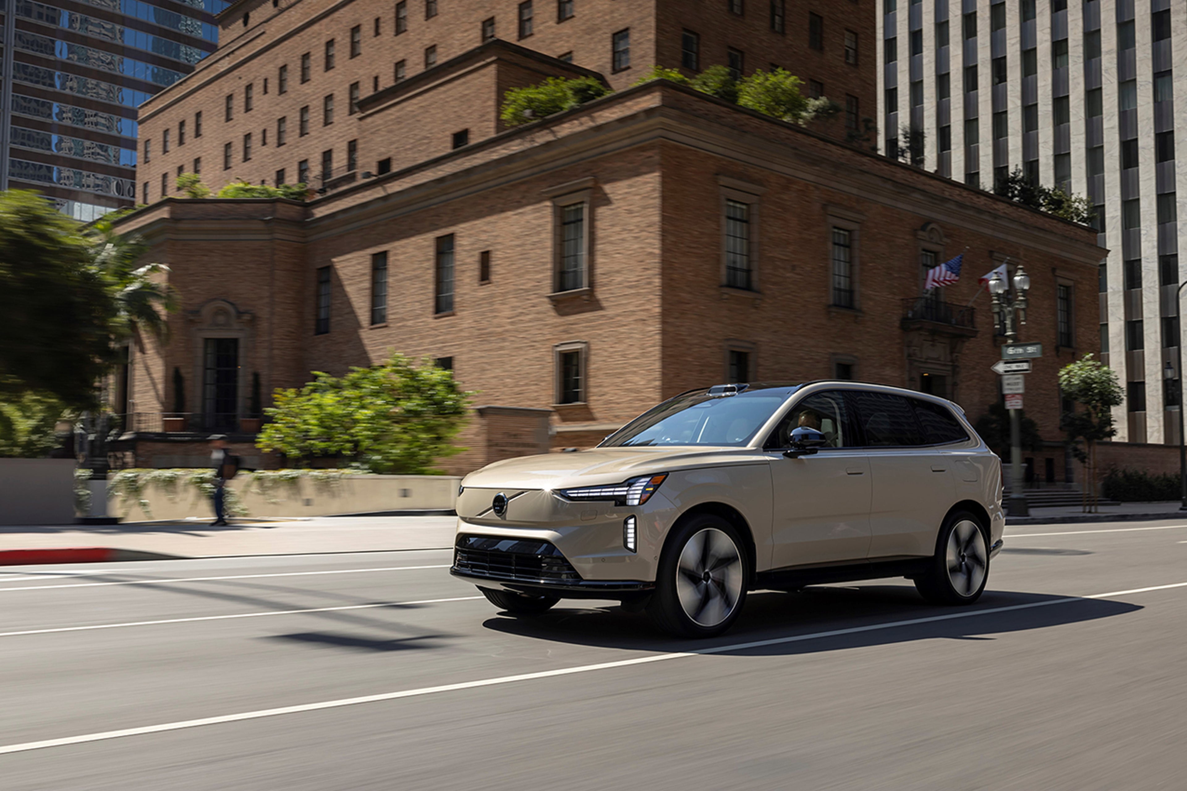 Volvo EX90 SUV me ngjyrë Sand Dune duke ecur në një rrugë të qytetit përpara një ndërtese historike me tulla.