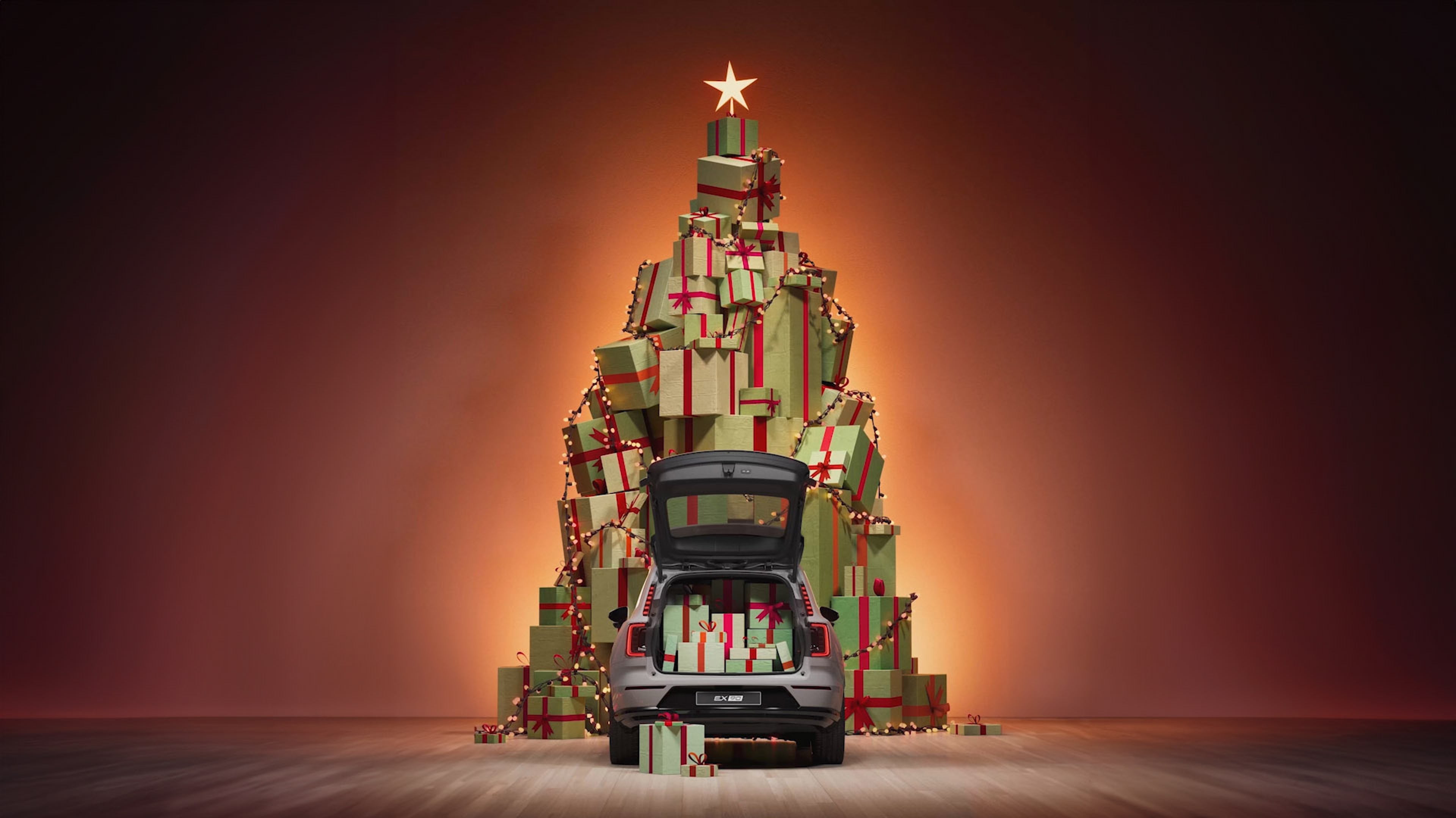 Фотографија на сив Volvo EX90 со багажник полн со подароци, паркиран пред новогодишна елка.