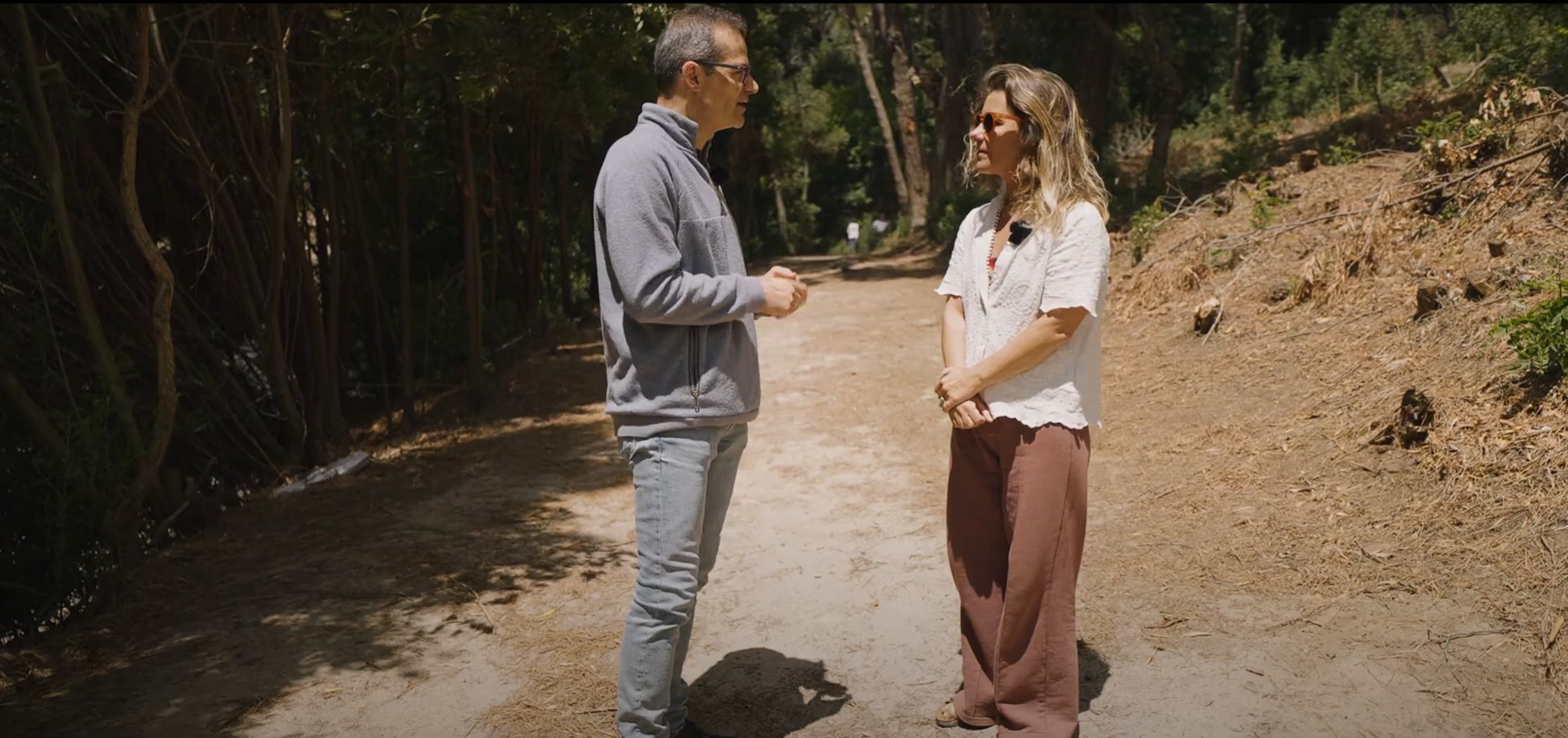 Duas pessoas conversam em um caminho de terra em meio a árvores. À esquerda, um homem de óculos veste um casaco cinza e calça jeans clara. À direita, uma mulher de óculos de sol veste uma blusa branca e calça larga marrom. O ambiente é ensolarado e rodeado por vegetação.