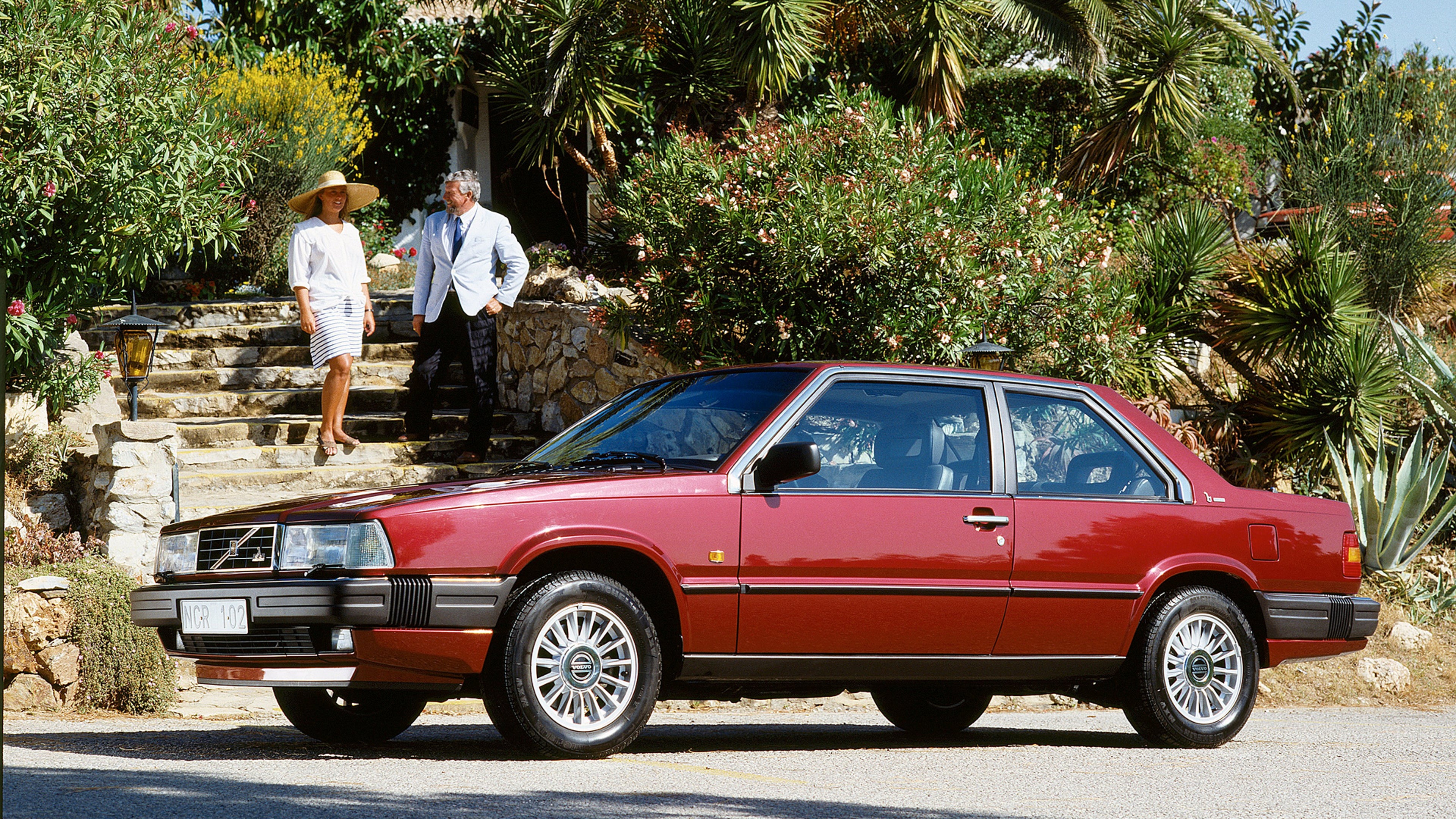 Classica Volvo 780 Bertone coupé di colore rosso, parcheggiata nel lussureggiante vialetto di un giardino con palme, due persone che conversano su gradini di pietra sotto un cielo azzurro e soleggiato.