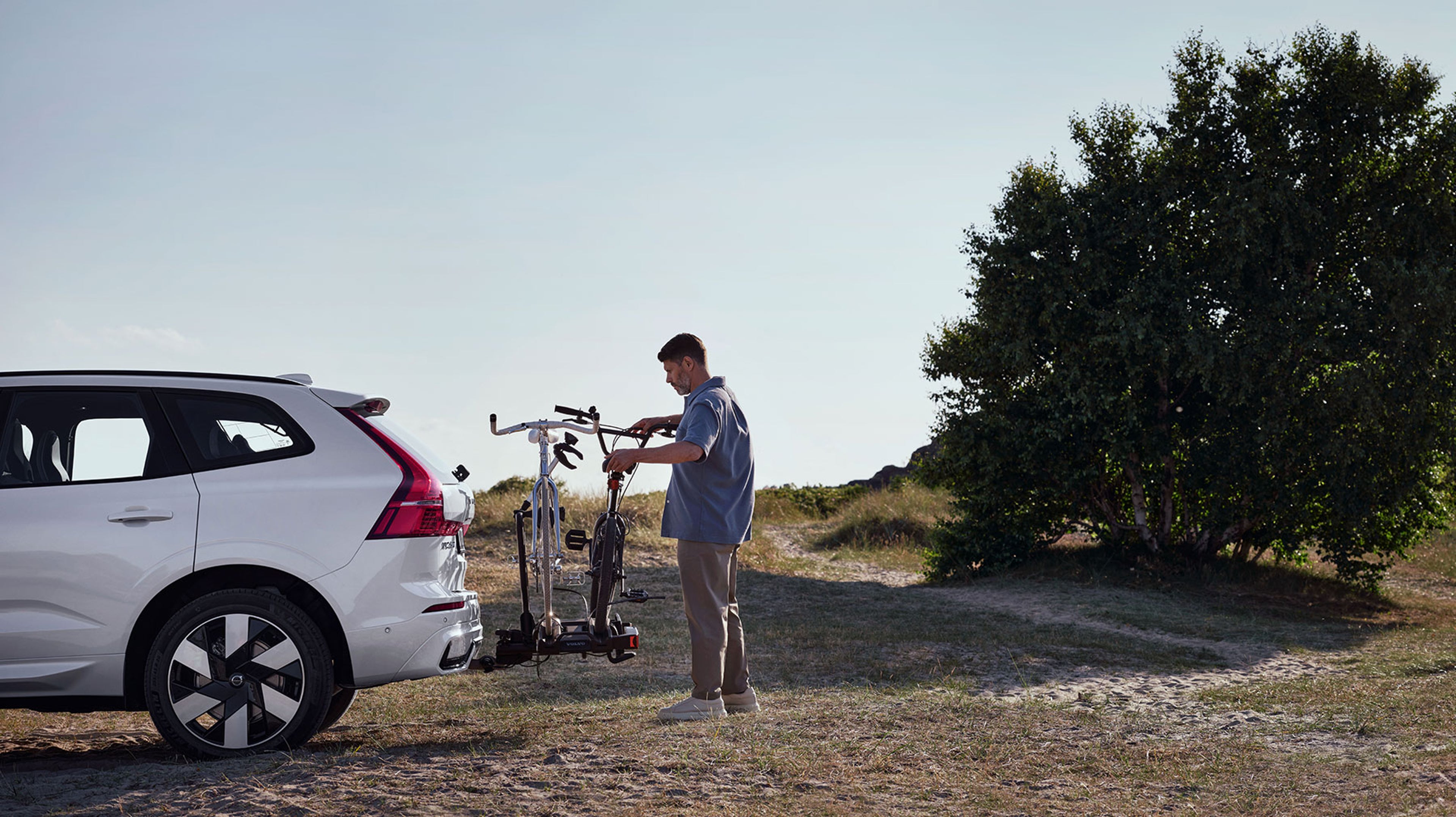 Een man zet zijn fiets op een opvouwbare fietsdrager op de trekhaak van een Volvo op een heidepad.