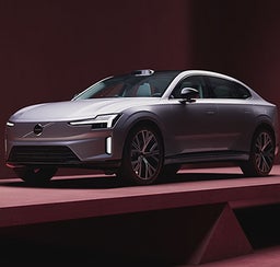 Front angle view of a VolvoES90 electric sedan on display with modern LED headlights and aerodynamic design, showcased in a minimalist studio with deep red lighting.