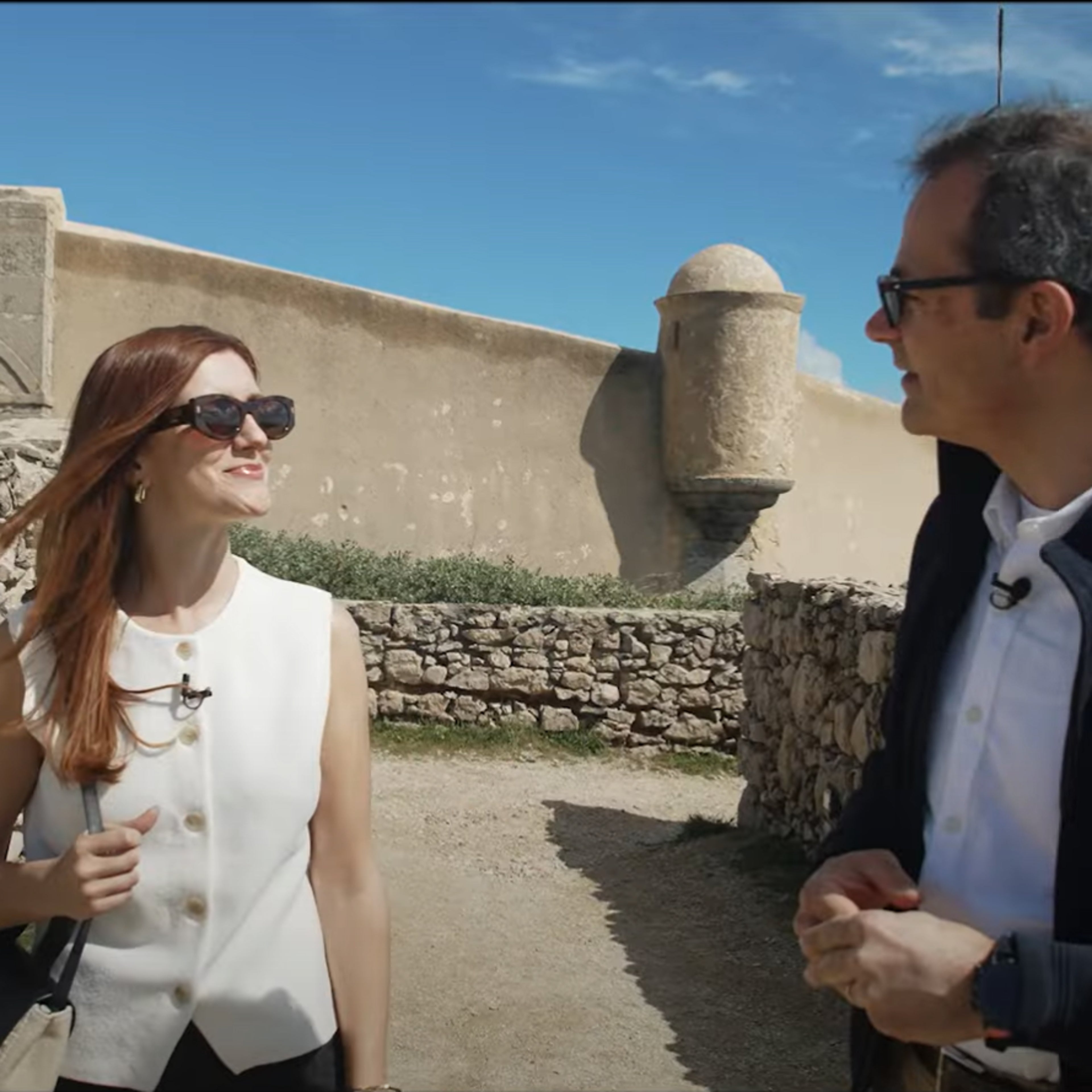 Duas pessoas, um homem e uma mulher, conversam ao ar livre junto a uma antiga estrutura de pedra, possivelmente uma fortaleza costeira. Ambos usam óculos de sol e microfones de lapela. Ao fundo, vê-se uma parede com um brasão de pedra e o mar sob um céu azul com algumas nuvens.