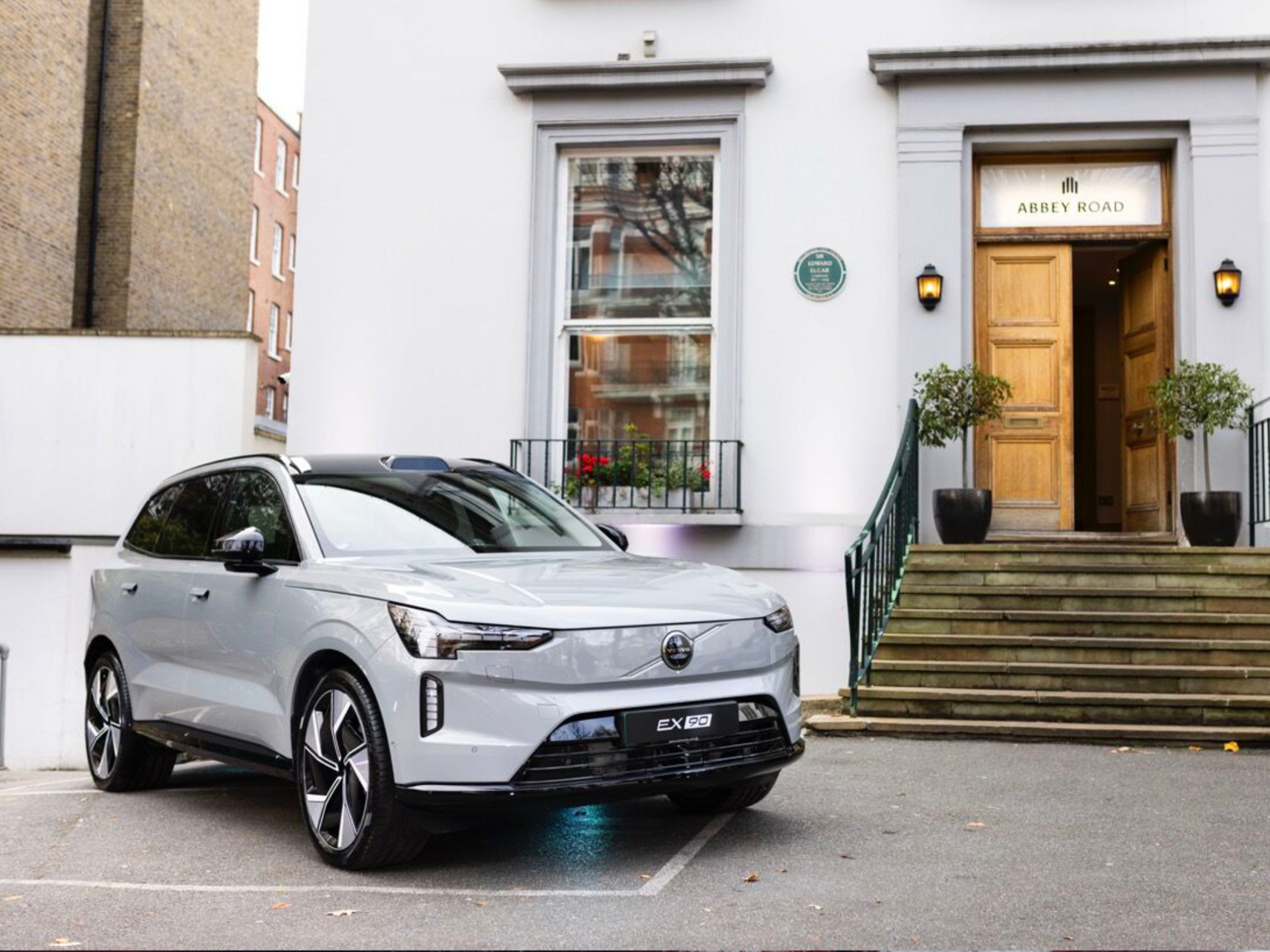 Ein grauer Volvo EX90 steht vor einem Haus mit geöffneter Tür über der "Abbey Road" steht.
